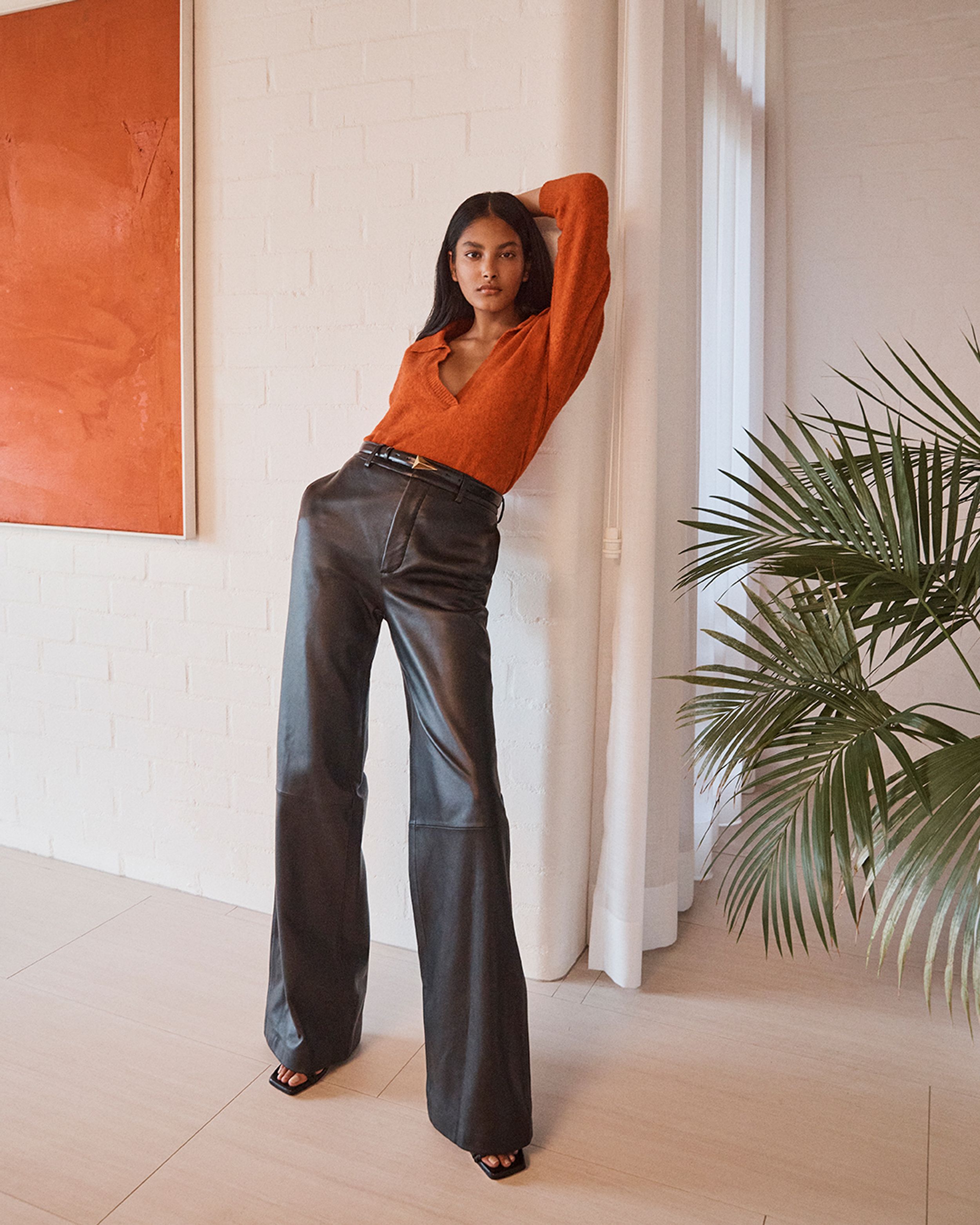 Brunette model leaning against a wall wearing an orange knit top and black leather trousers