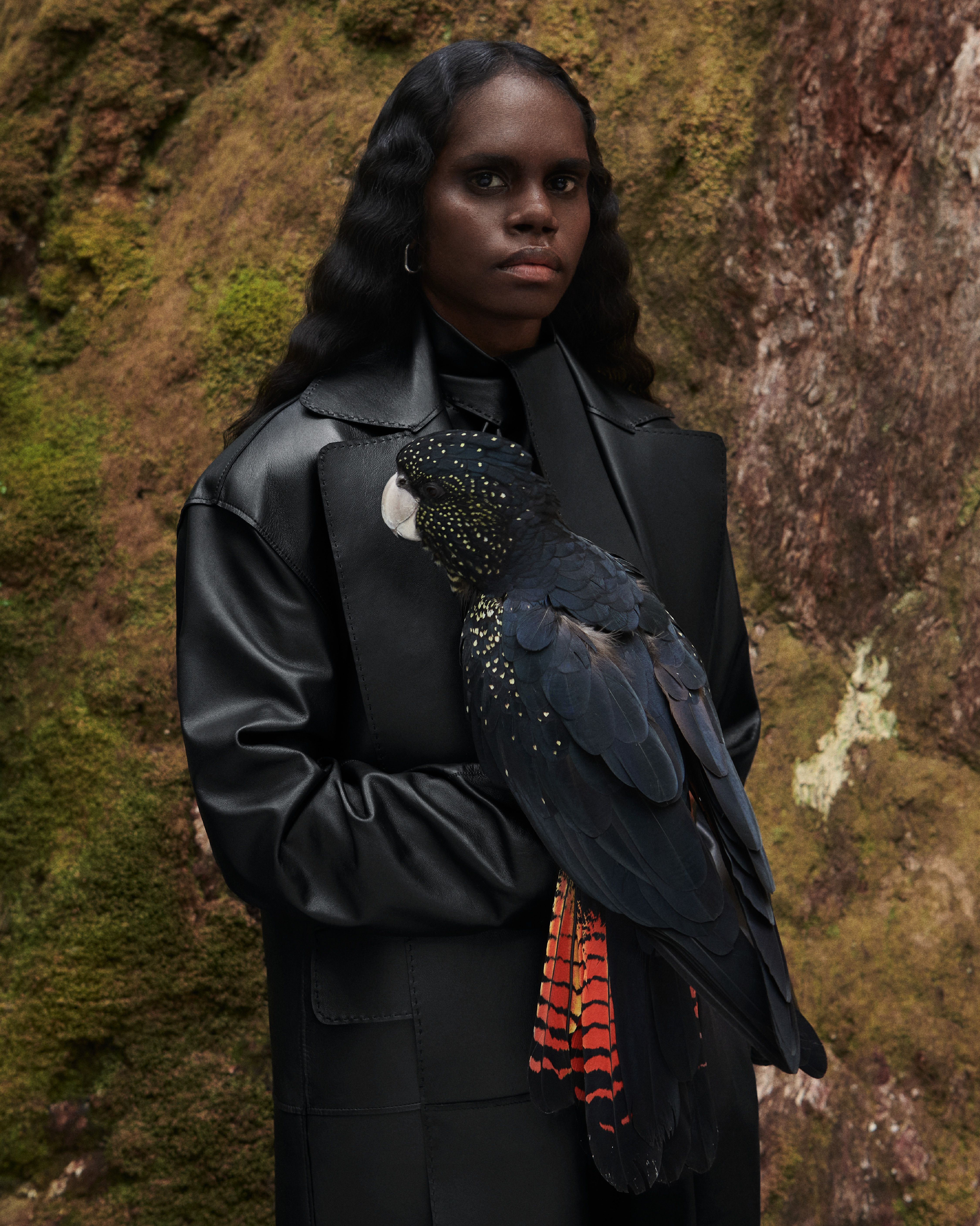Model Tarlisa wearing a black leather top and trench coat with a cockatoo standing in front of a tree.
