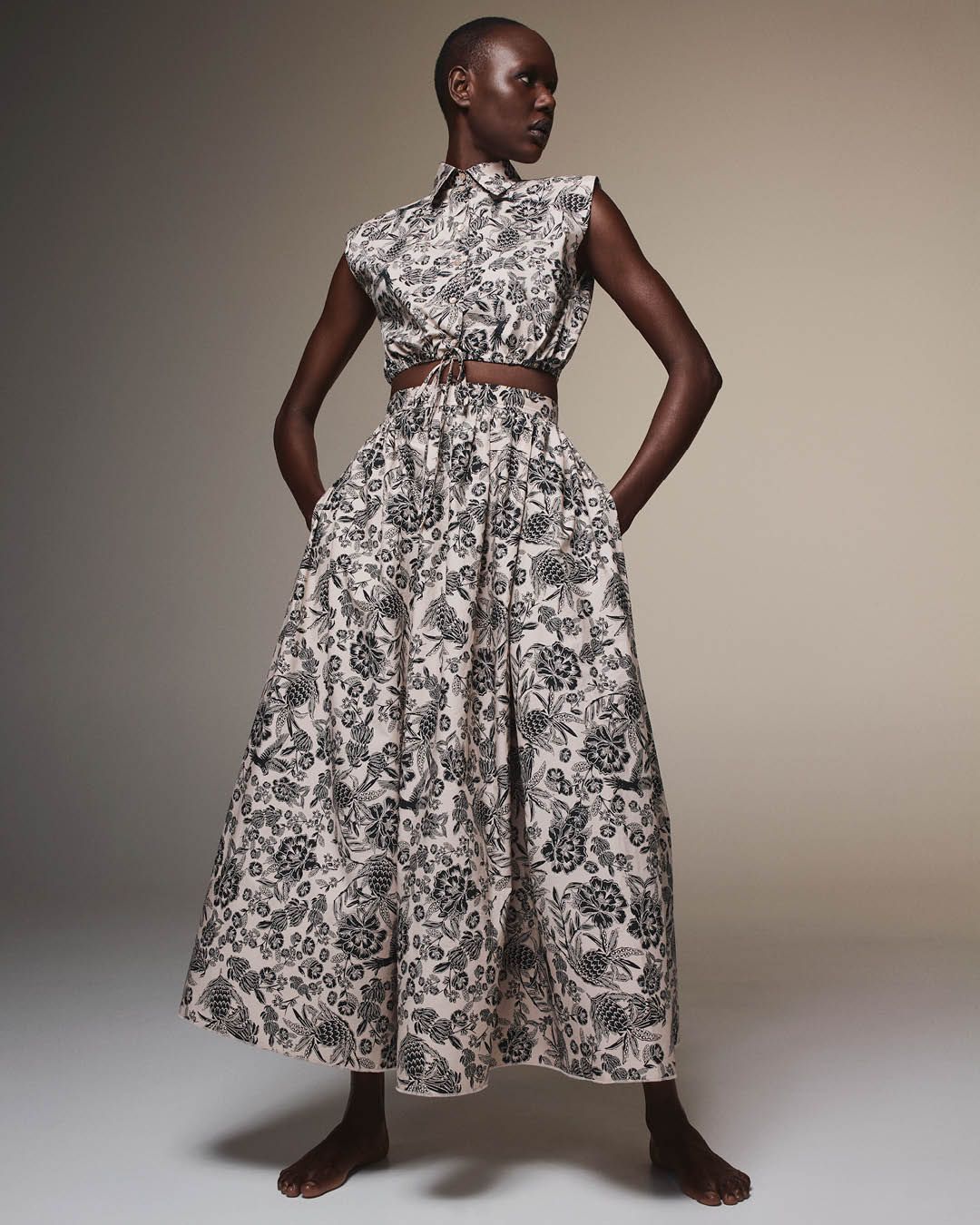 Ajak Deng standing with hands in pockets and head turned wearing a matching grey floral cropped shirt and long skirt