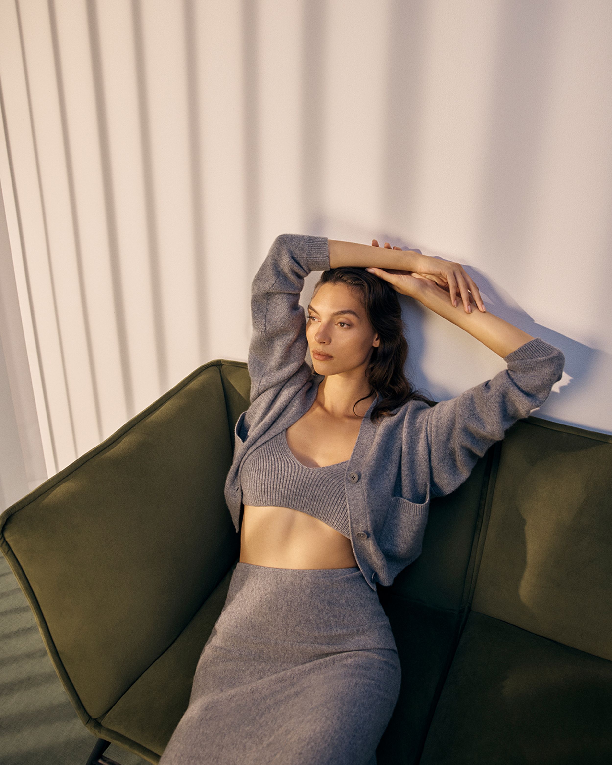 Brunette model sitting on a green sofa with both arms on her head wearing a grey cashmere singlet, cardigan and skirt