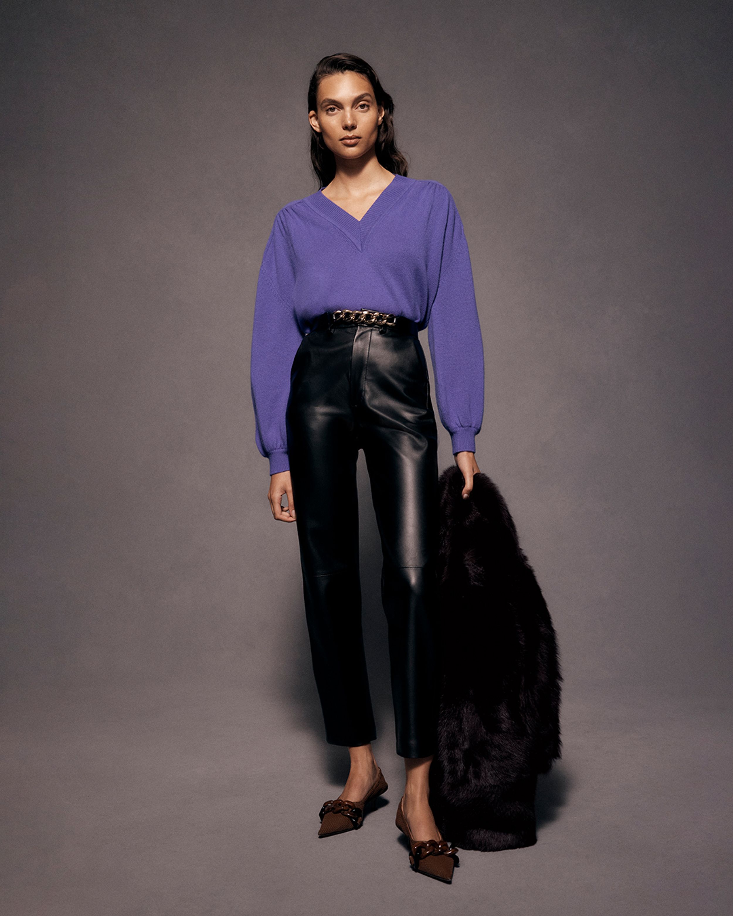 Brunette model standing in a purple v-neck sweater and leather trousers, holding a black jacket