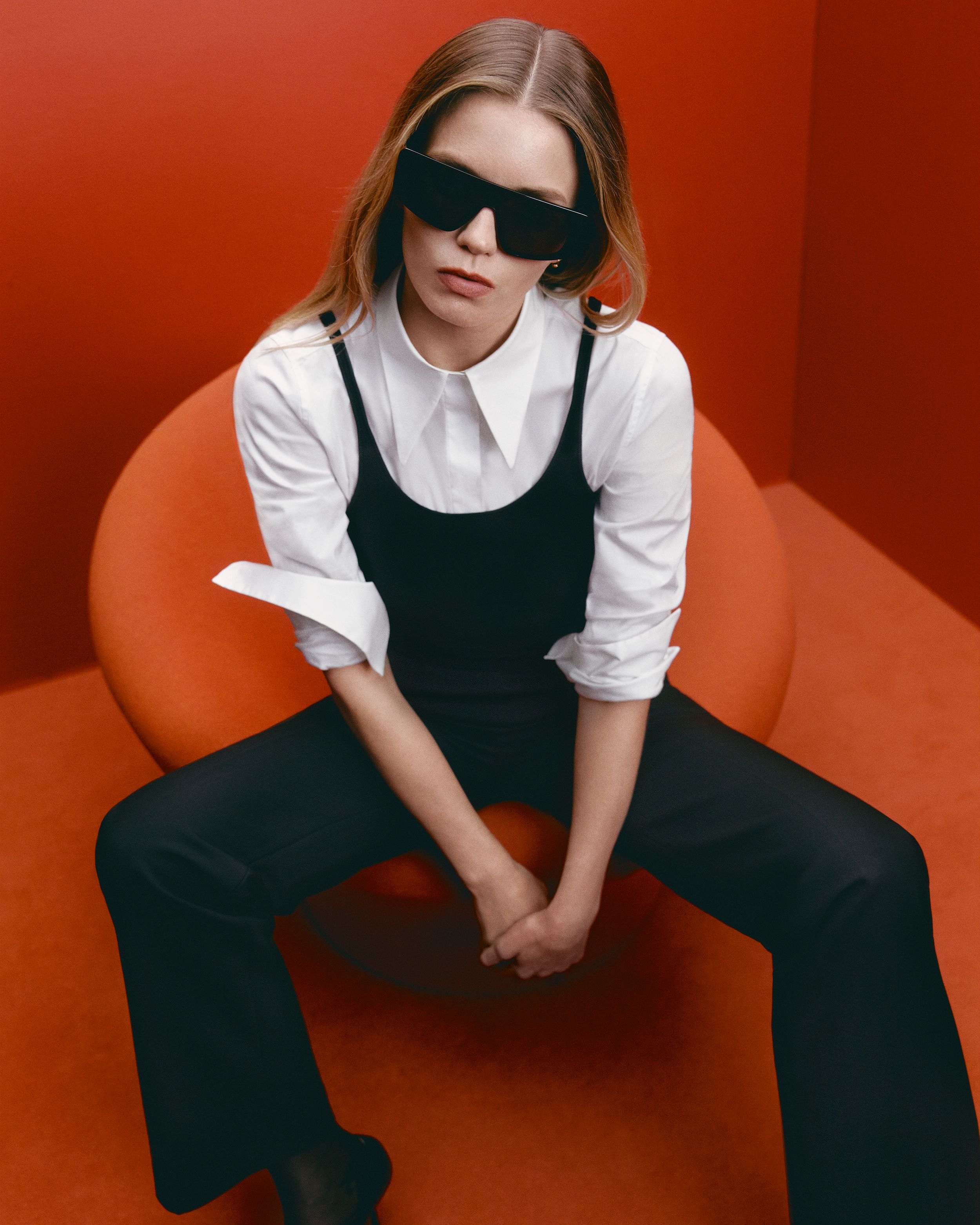 Abbey Lee sitting on a red chair in a red room wearing a white shirt, black camisole, trousers and sunglasses