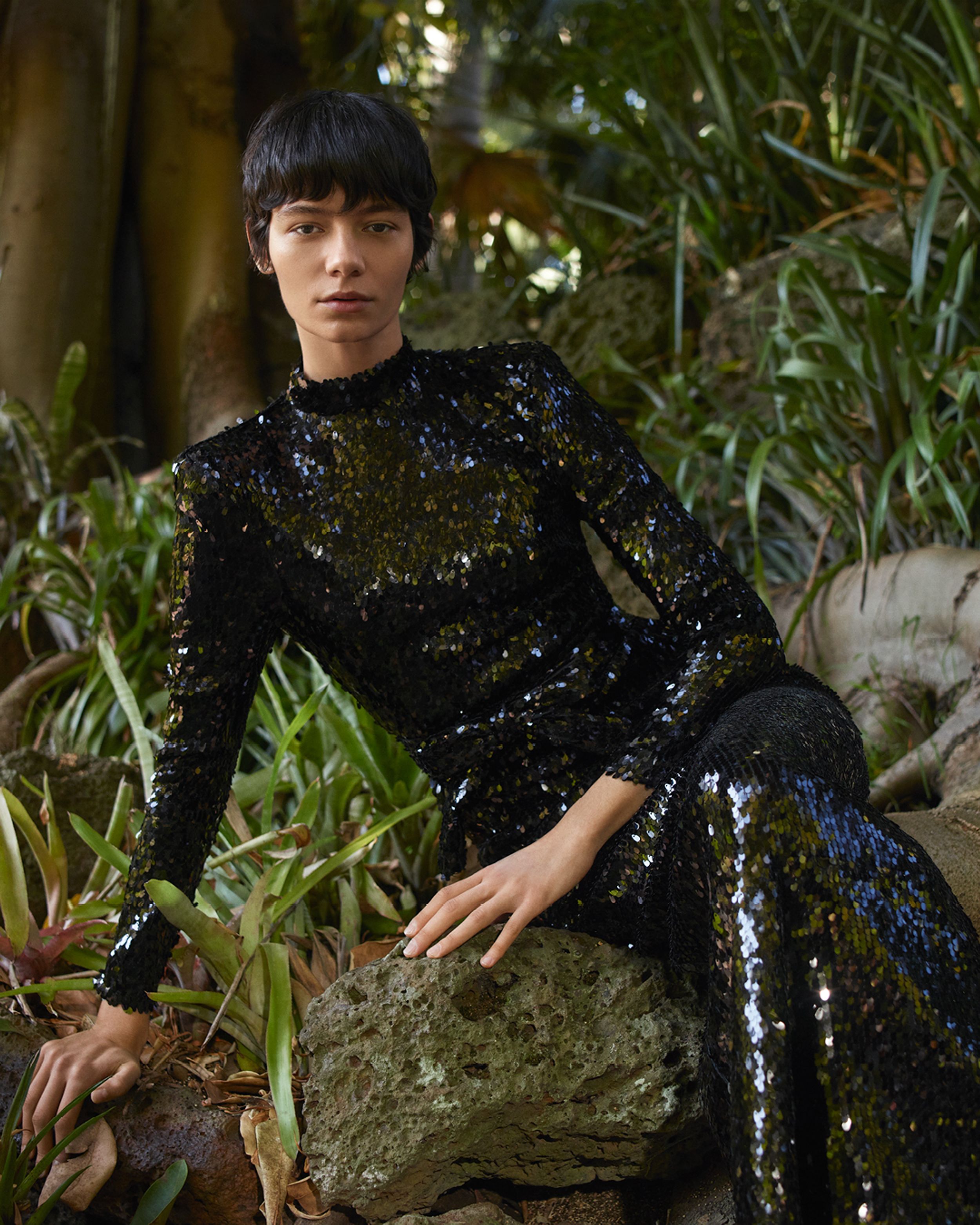 Model perched on a rock amongst greenery wearing a black sequined gown