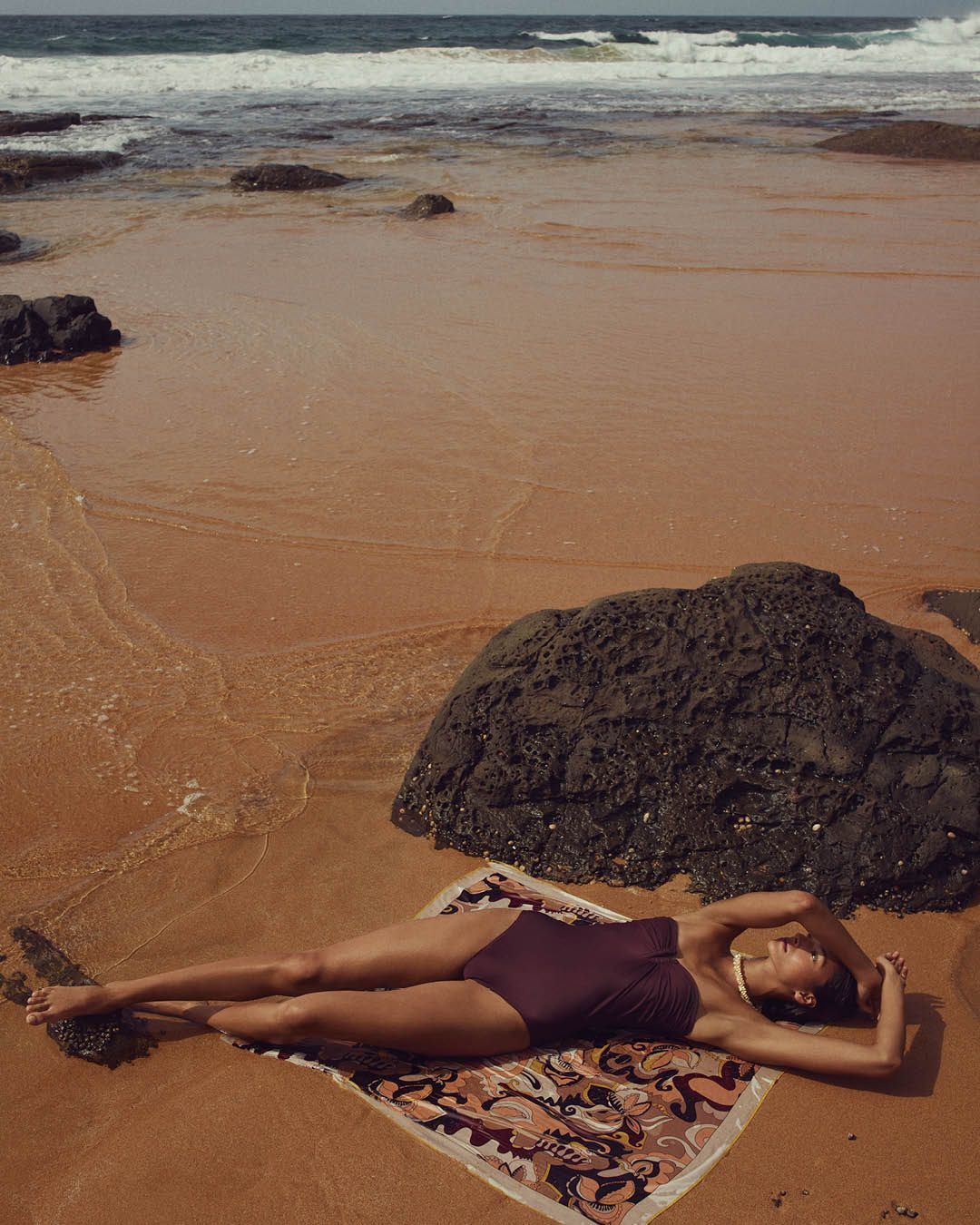 Model laying on a neutral-patterned scarf on a sandy beach wearing a brown one-piece swimsuit