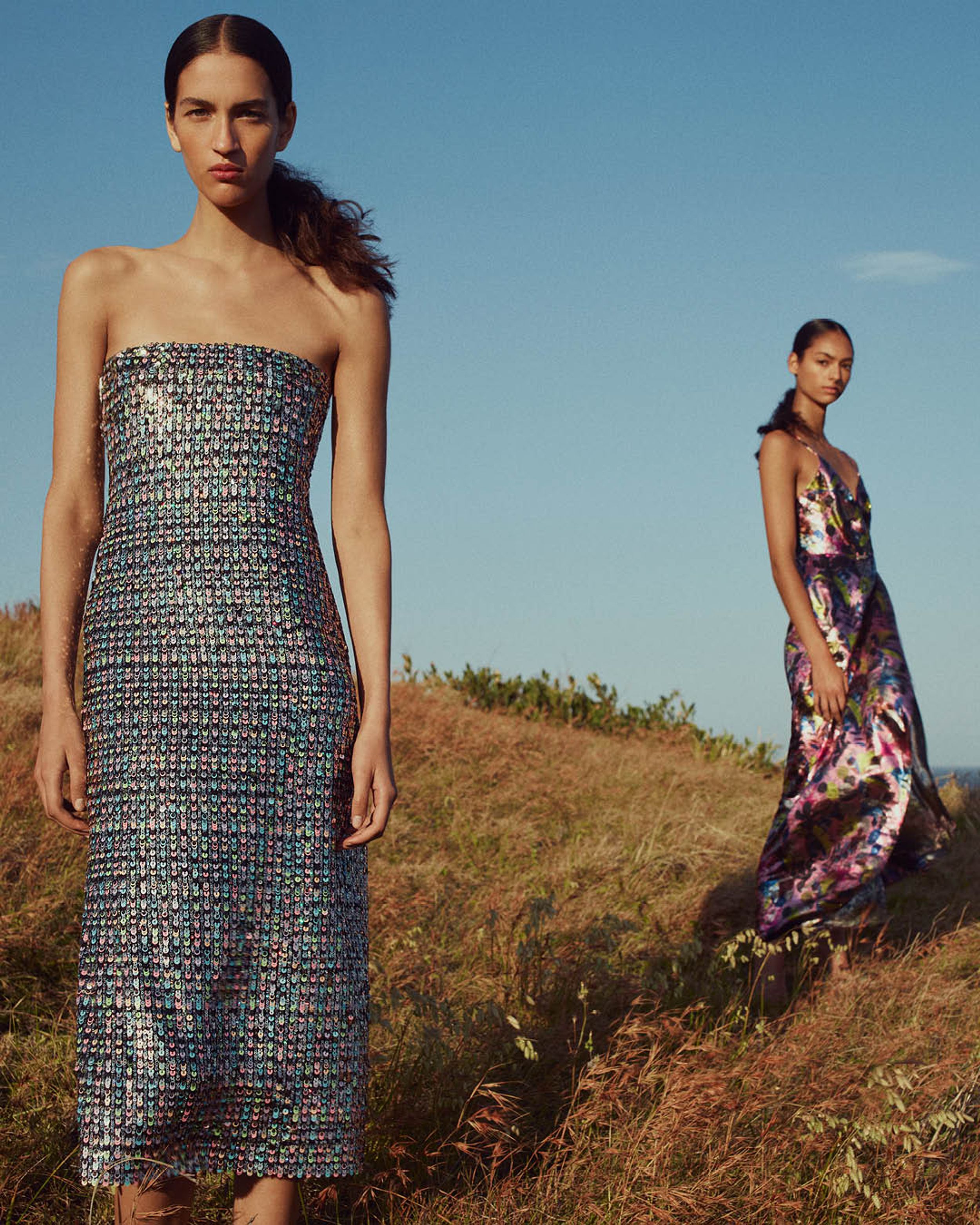 Two models standing on grass gazing at the camera wearing multicoloured shimmering evening gowns