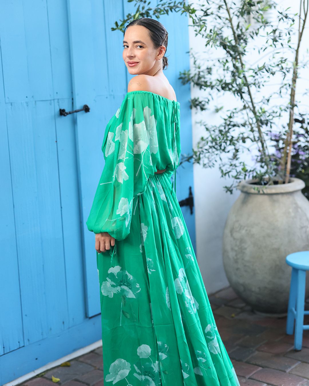 Woman looking over shoulder at camera wearing off-shoulder green floral print dress in front of blue door
