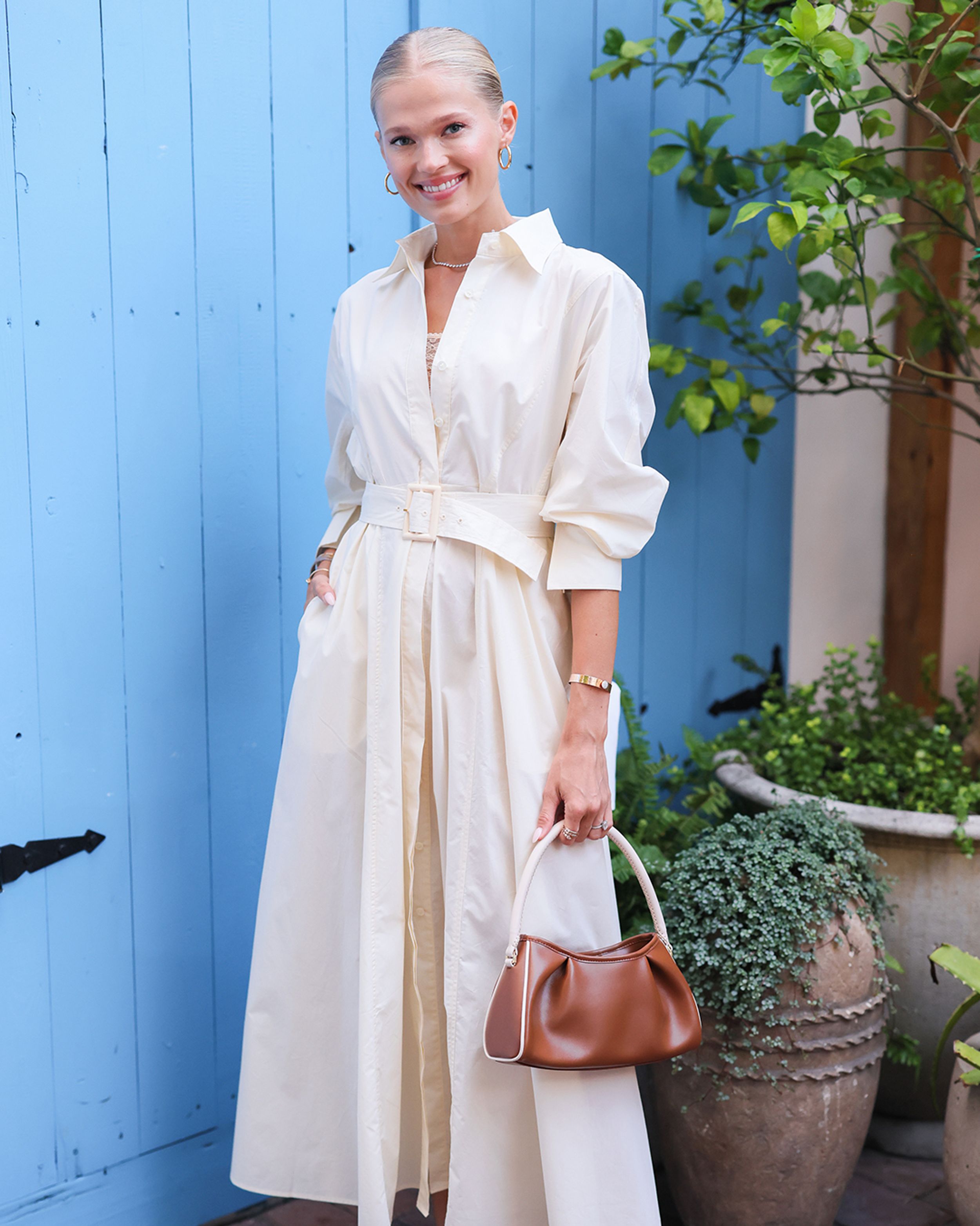 Blonde woman smiling wearing light yellow shirt dress with one hand in pocket and brown bag in hand