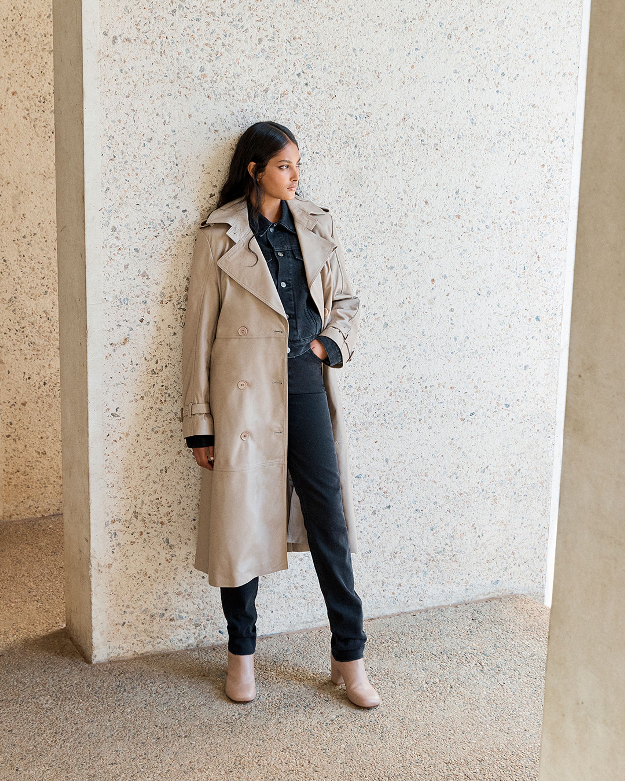 Brunette model leaning against a wall wearing a neutral leather trench, denim shirt and jeans