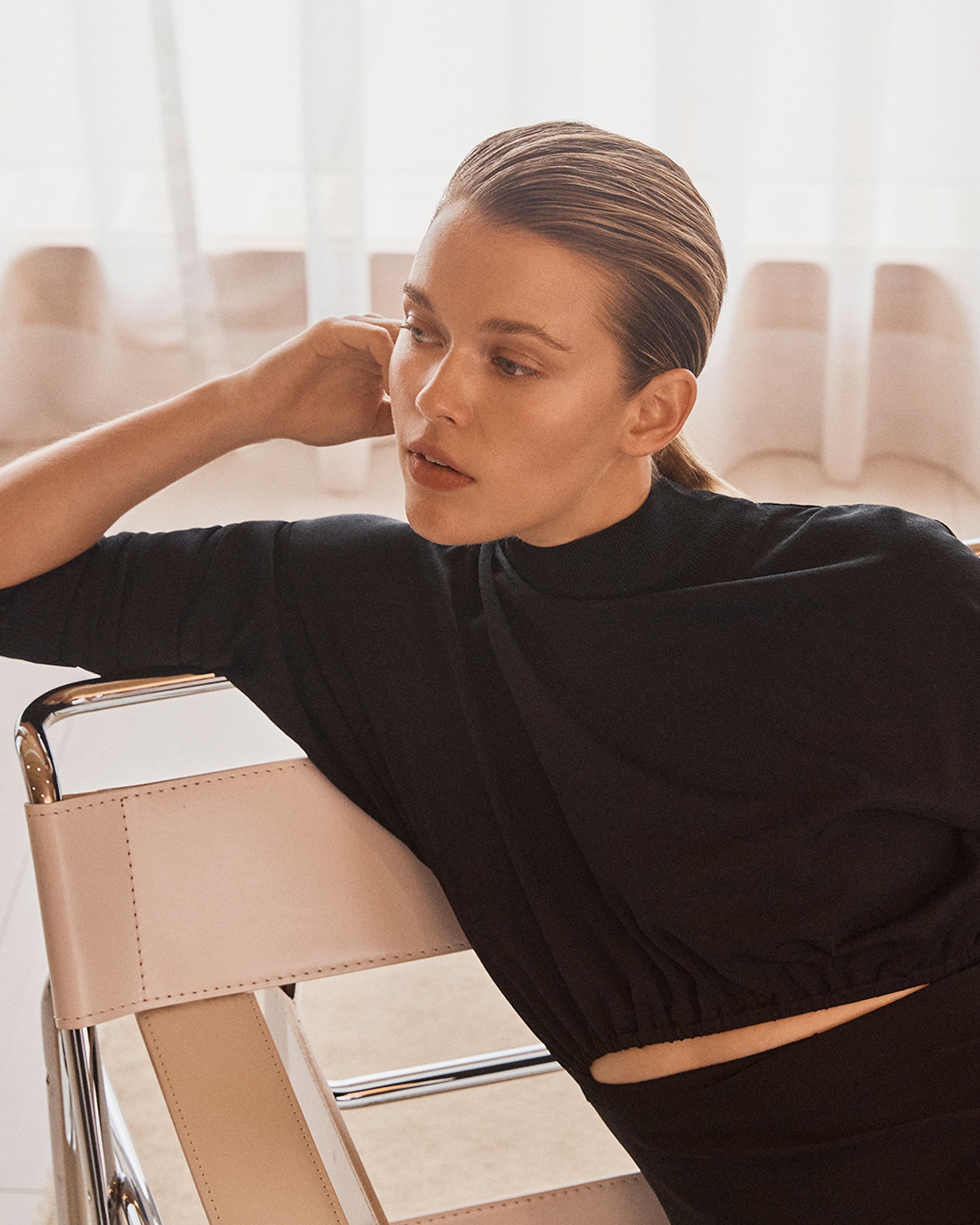 Model leaning on armchair looking to the side wearing a black long sleeve dress