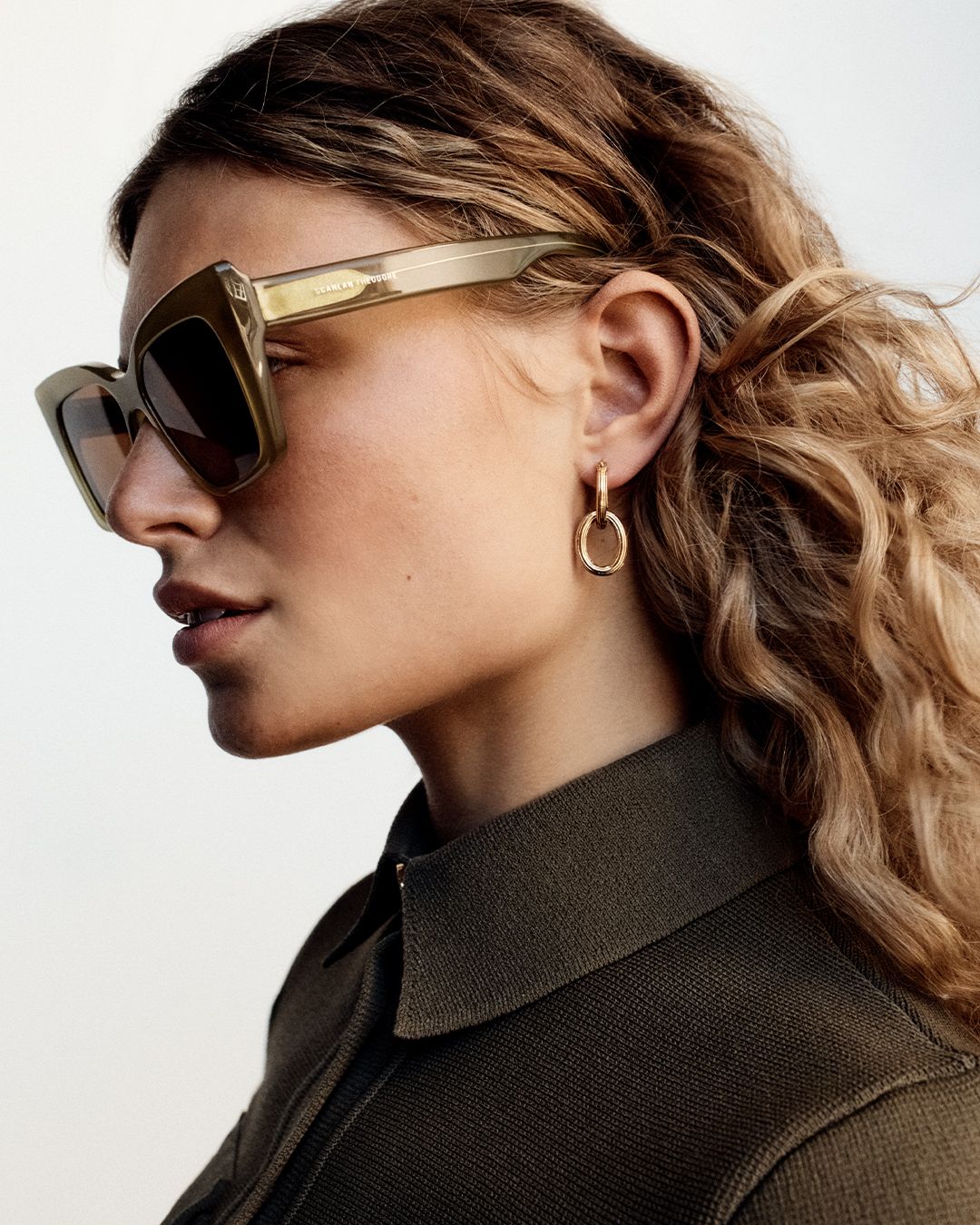 Model looking to the side wearing brown sunglasses and a khaki collared Crepe Knit dress with long curly blonde hair