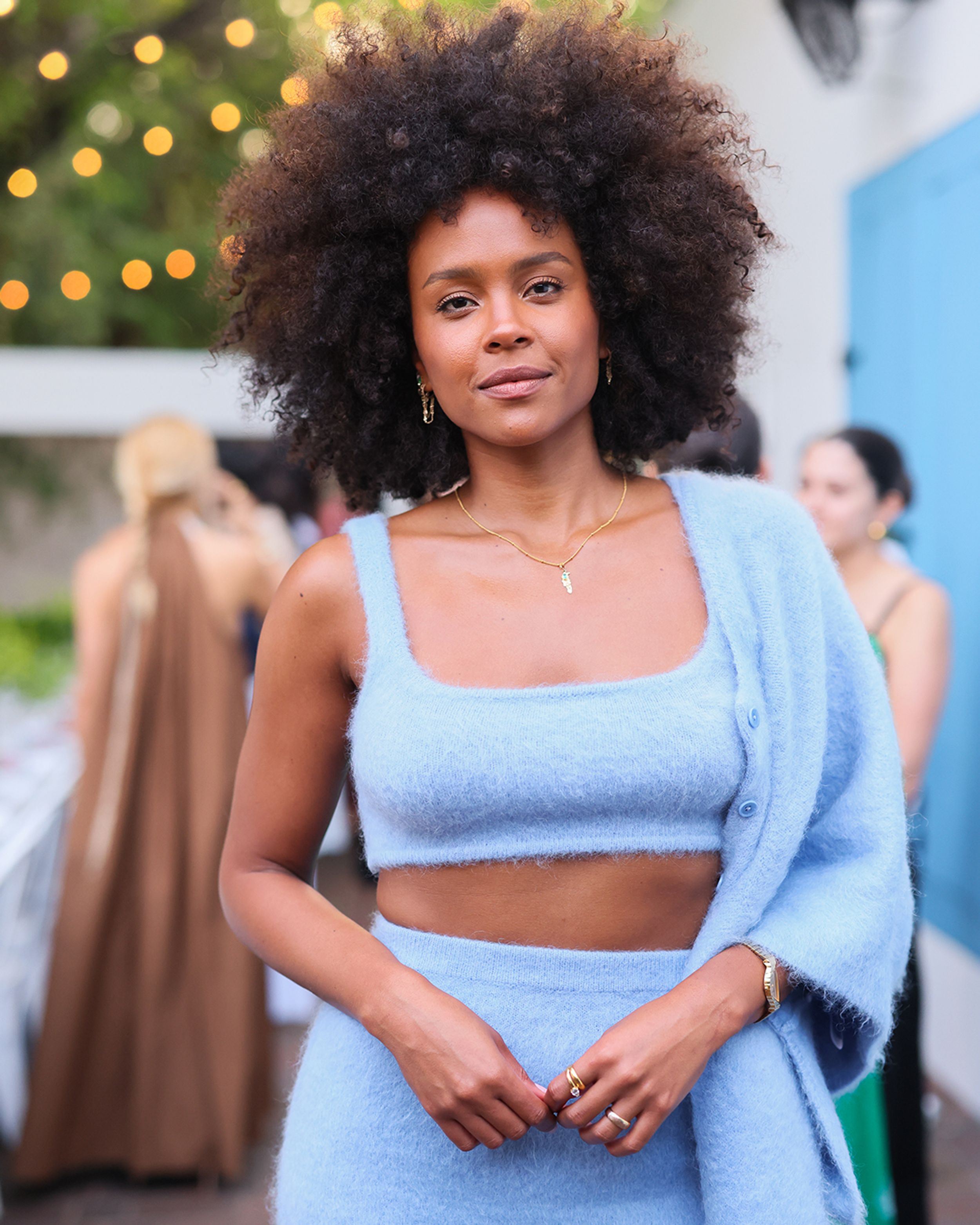 Woman with brown hair smiling hands crossed wearing blue knitted cardigan, crop singlet and skirt set