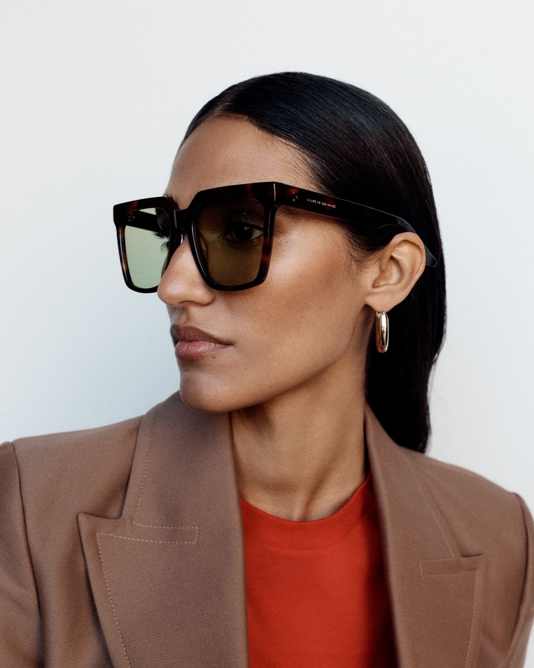 Model looking to the side wearing brown oversized sunglasses with a brown blazer and red top