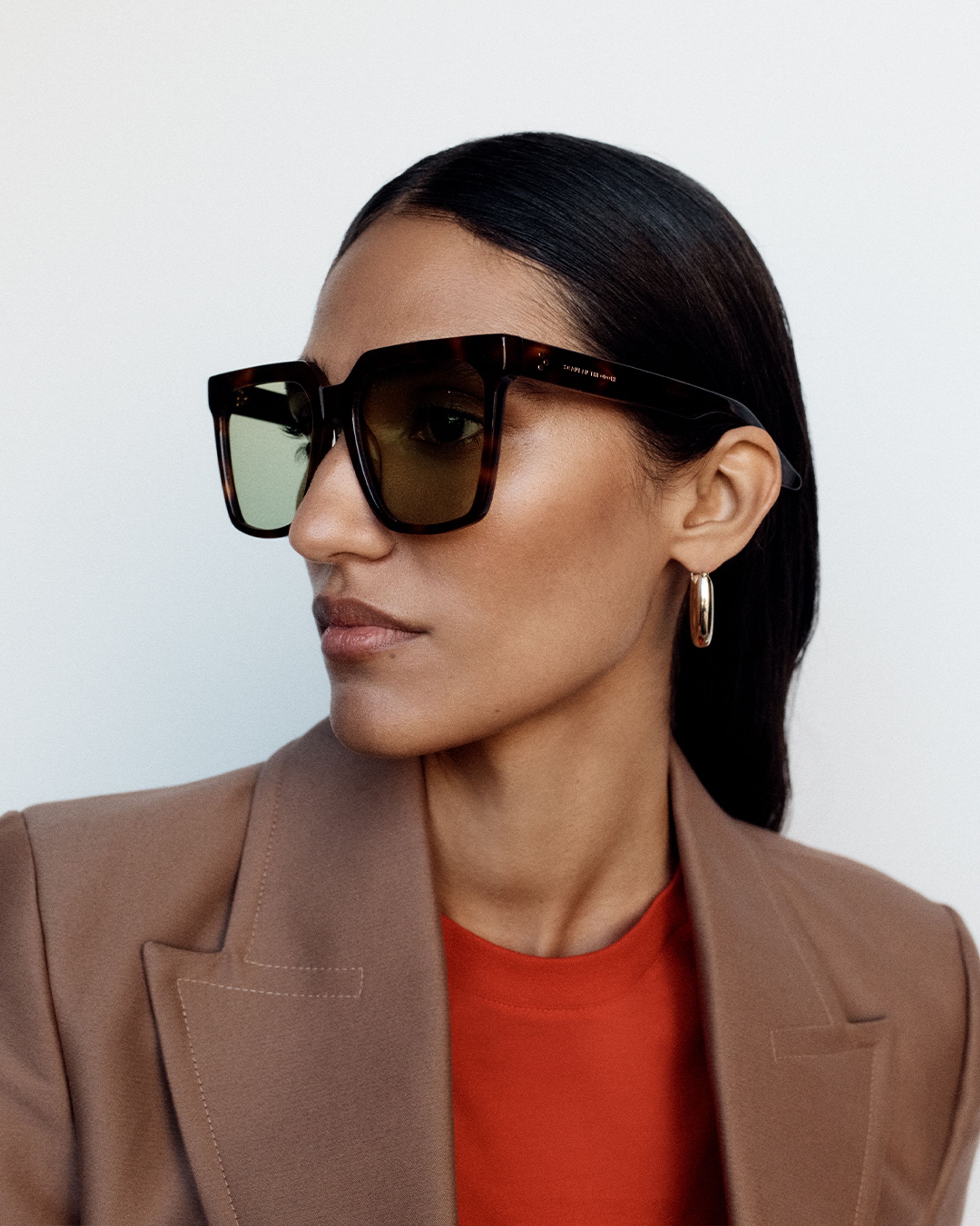 Model looking to the side wearing brown oversized sunglasses with a brown blazer and red top