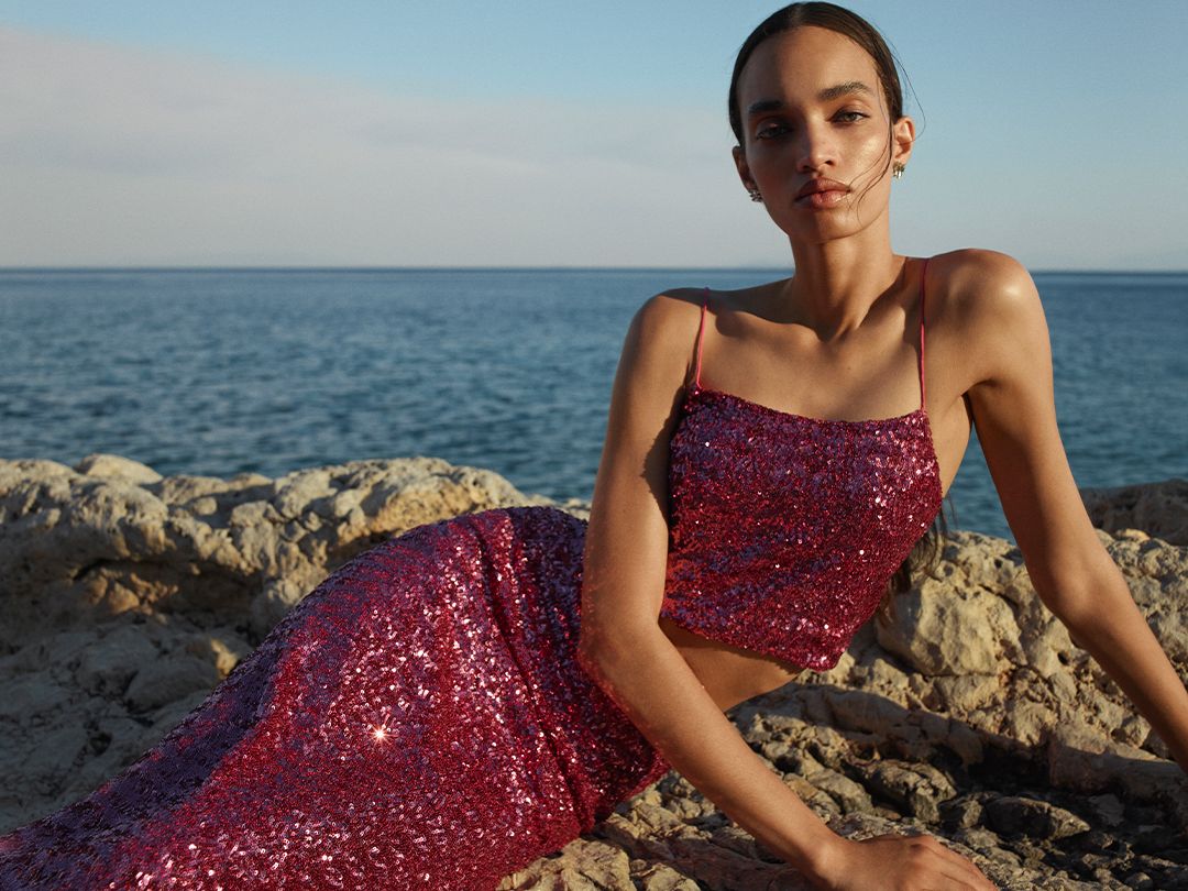 Model wearing a pink sequined singlet and skirt sitting on rocks with the ocean in the background