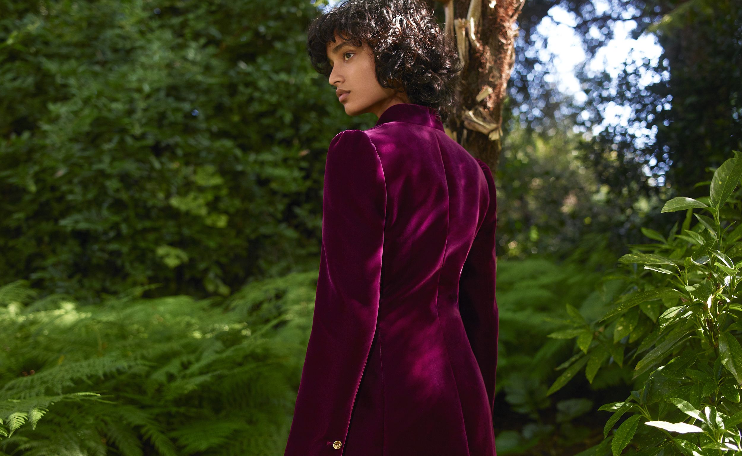 Models standing with her back turned amongst greenery wearing a mulberry velvet suit jacket