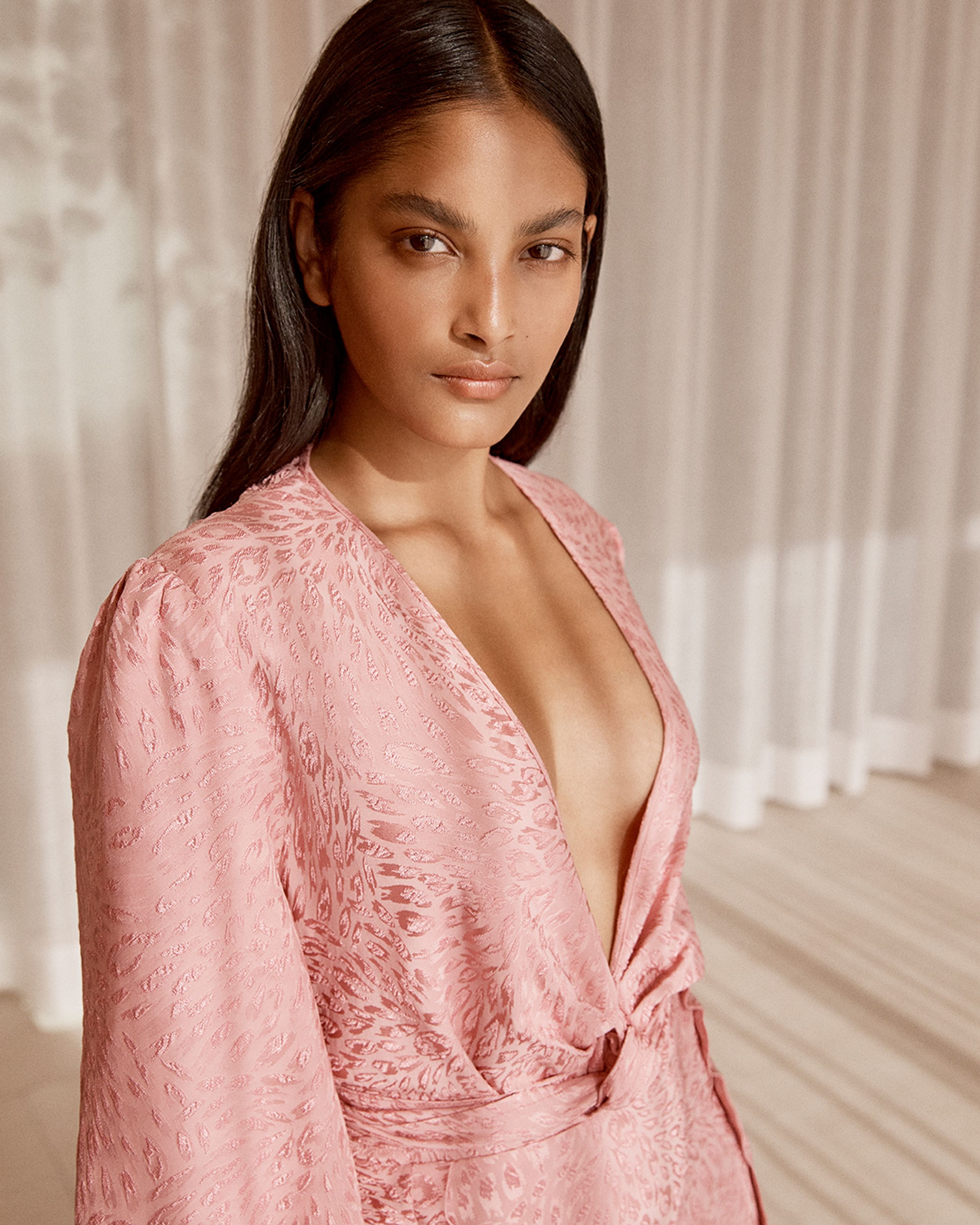 Brunette model standing by window drapes wearing a pink silk top