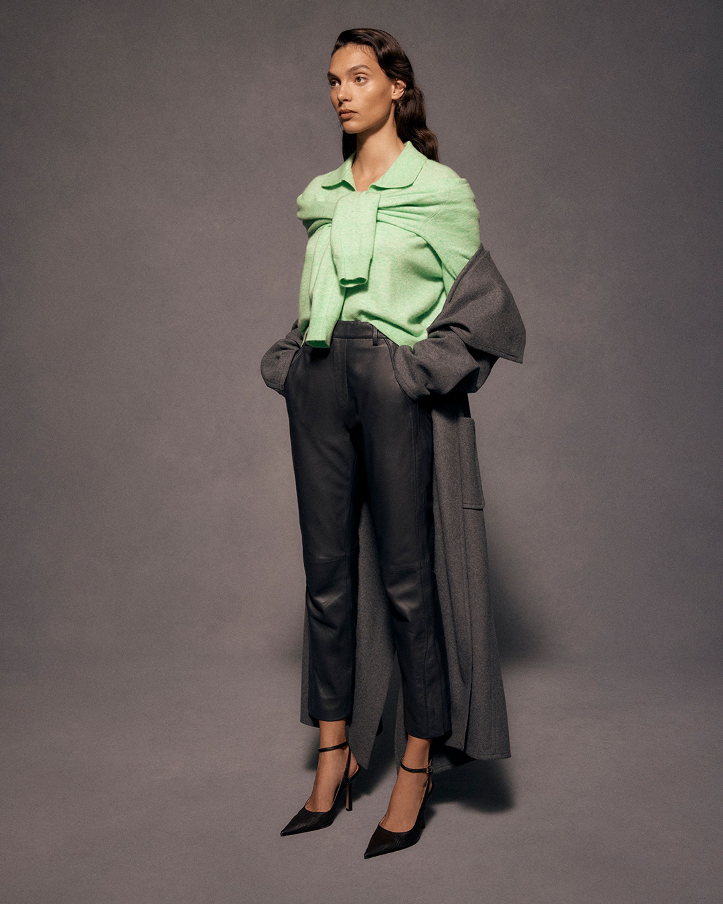 Brunette model facing the side, standing in a green knit and black trousers