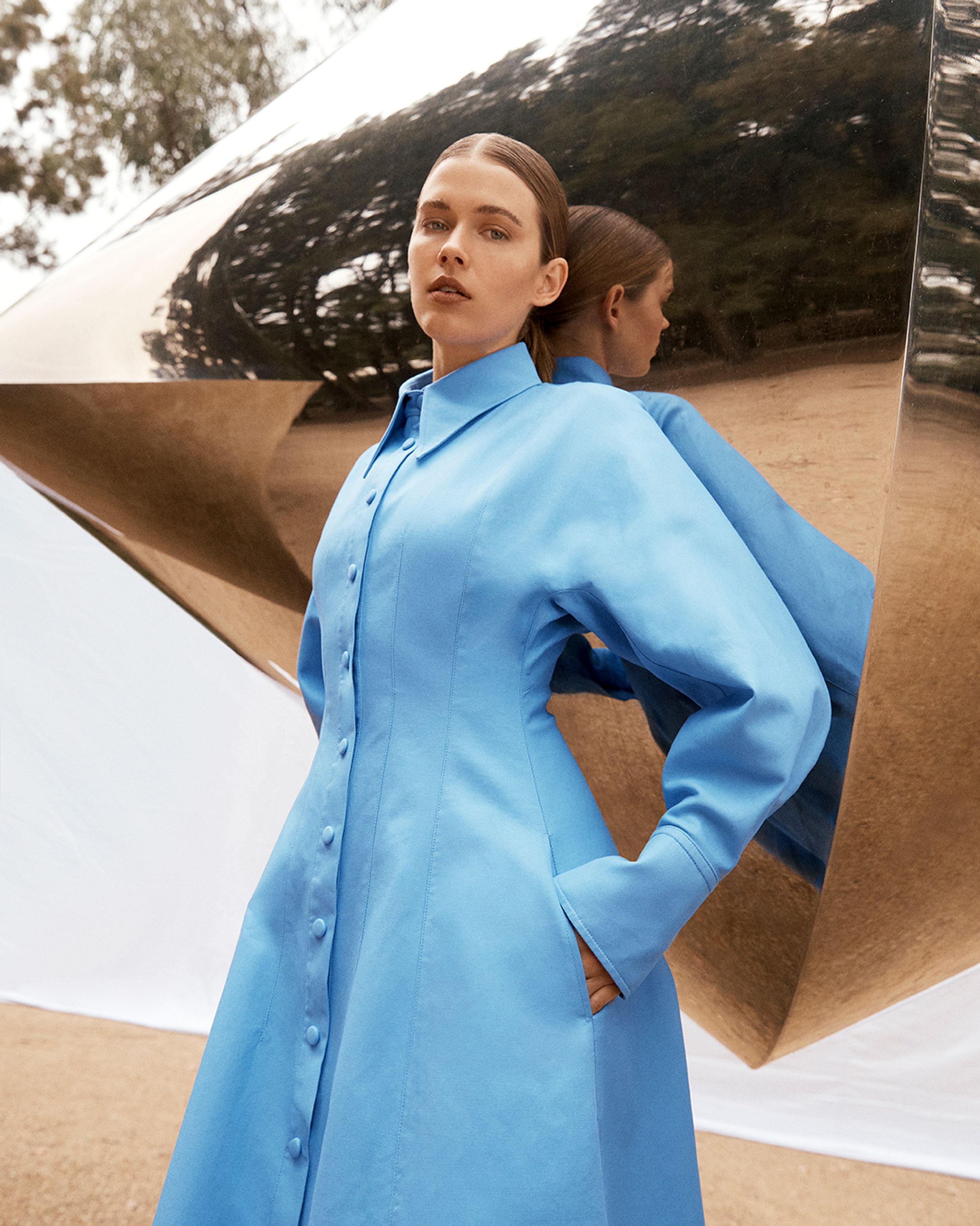 Brunette model leaning against mirrored sculpture wearing a blue shirt dress