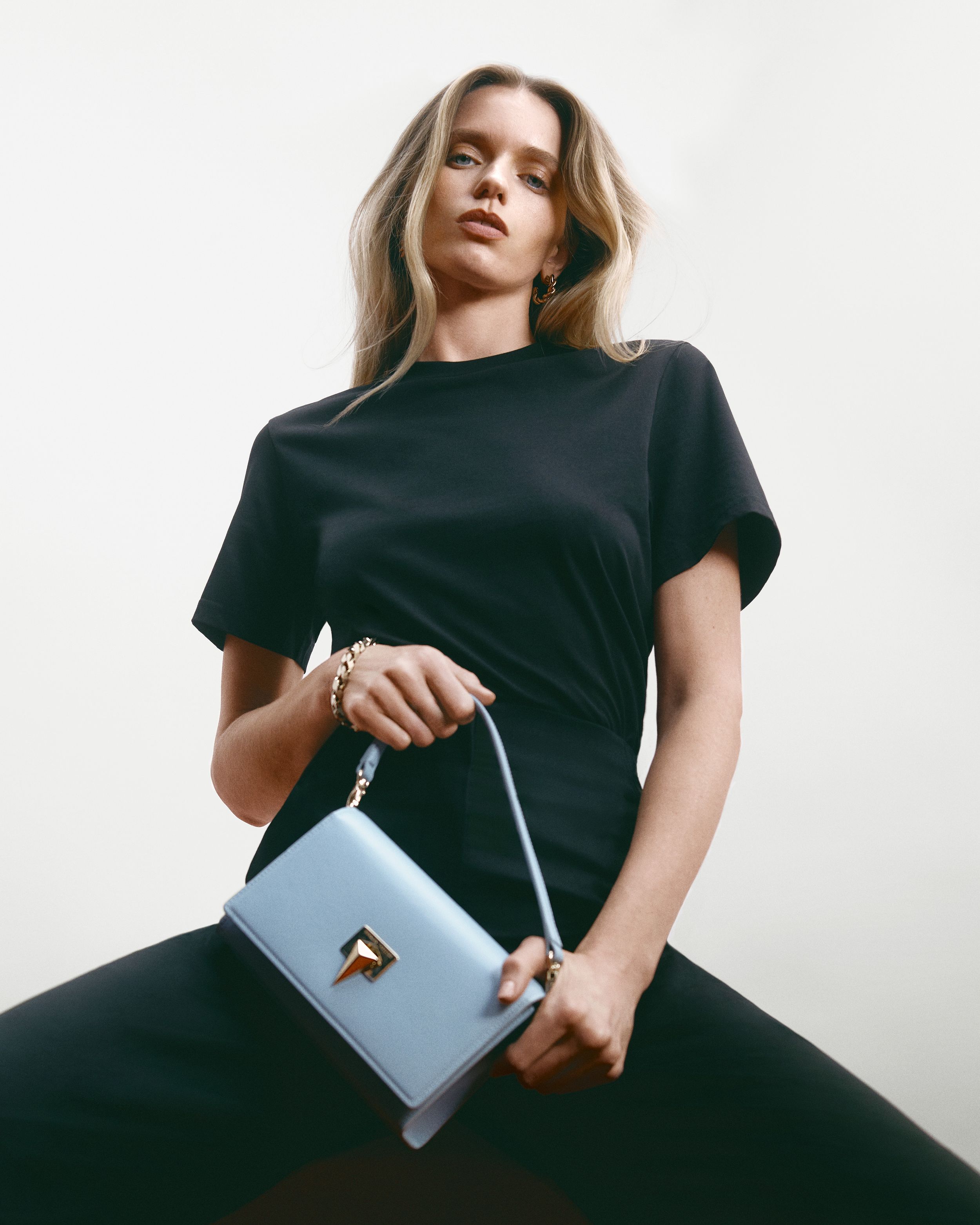 Abbey Lee sitting wearing a black tee shirt and trousers, carrying a baby blue shoulder bag