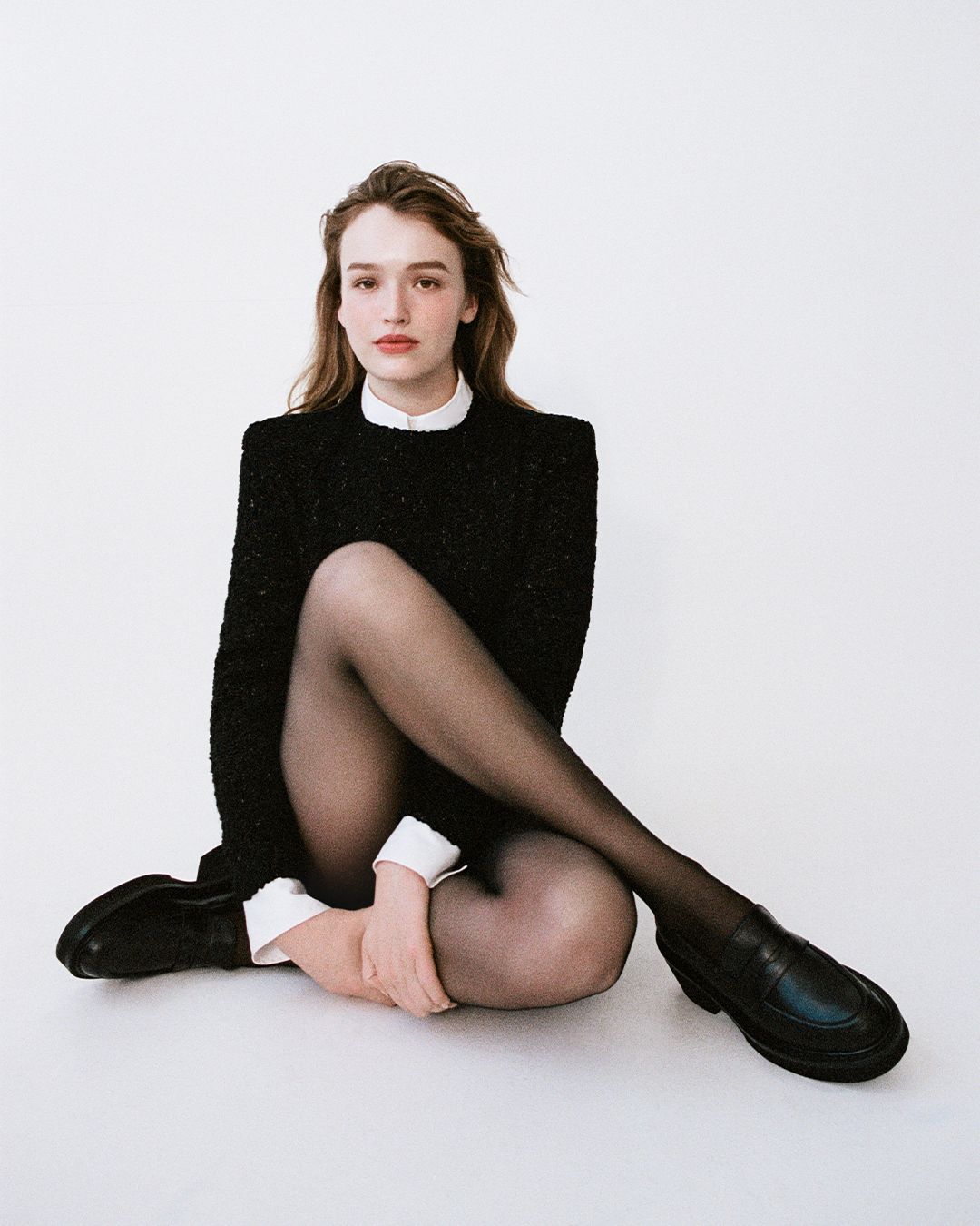 Maddison Brown sitting with legs crossed on the floor in black knitted top and white collared shirt