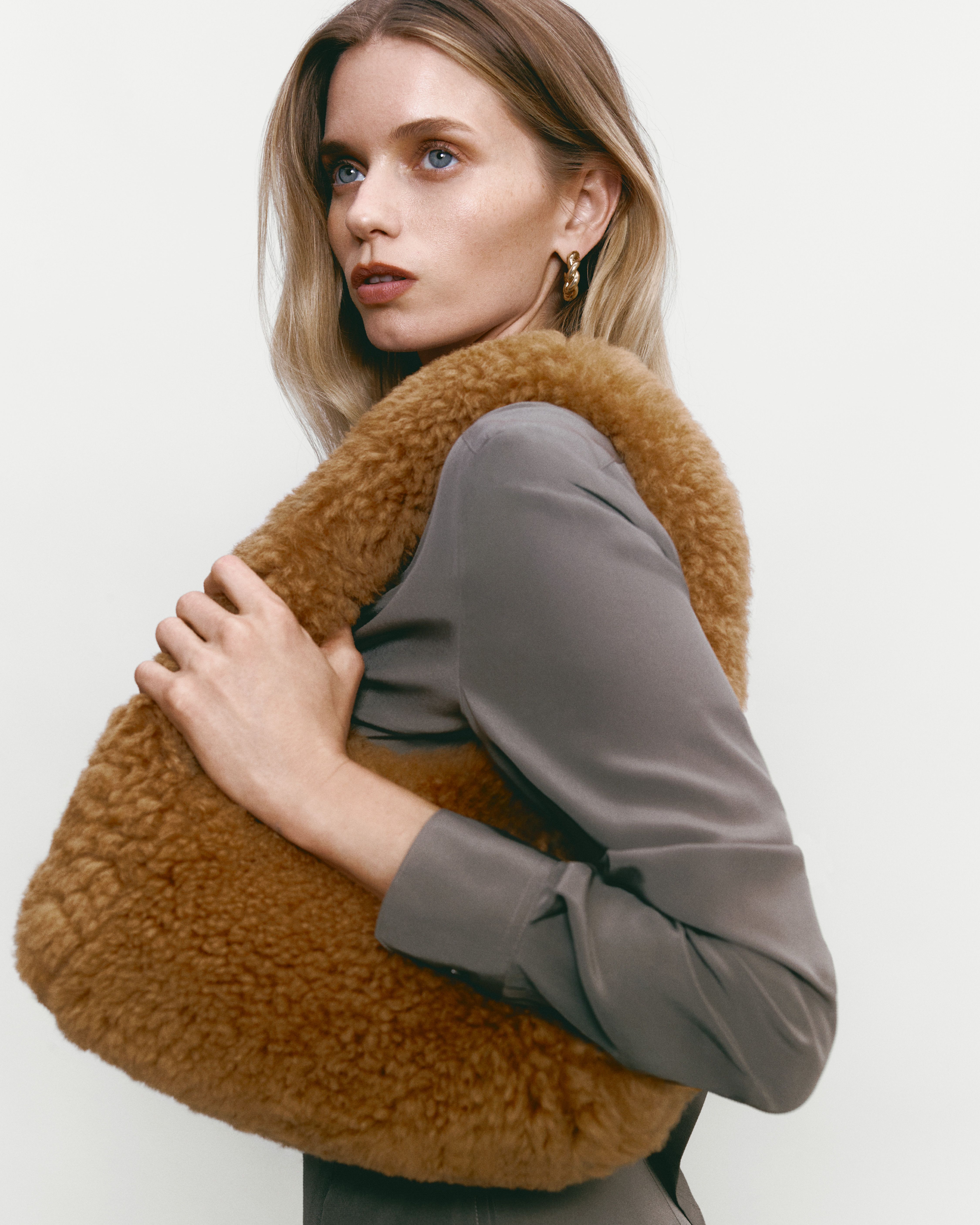 Abbey Lee facing the side wearing a taupe silk shirt with tan shearling bag on her shoulder