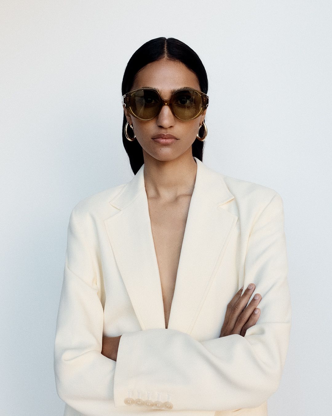 Model posing with arms crossed wearing khaki round sunglasses with a cream blazer