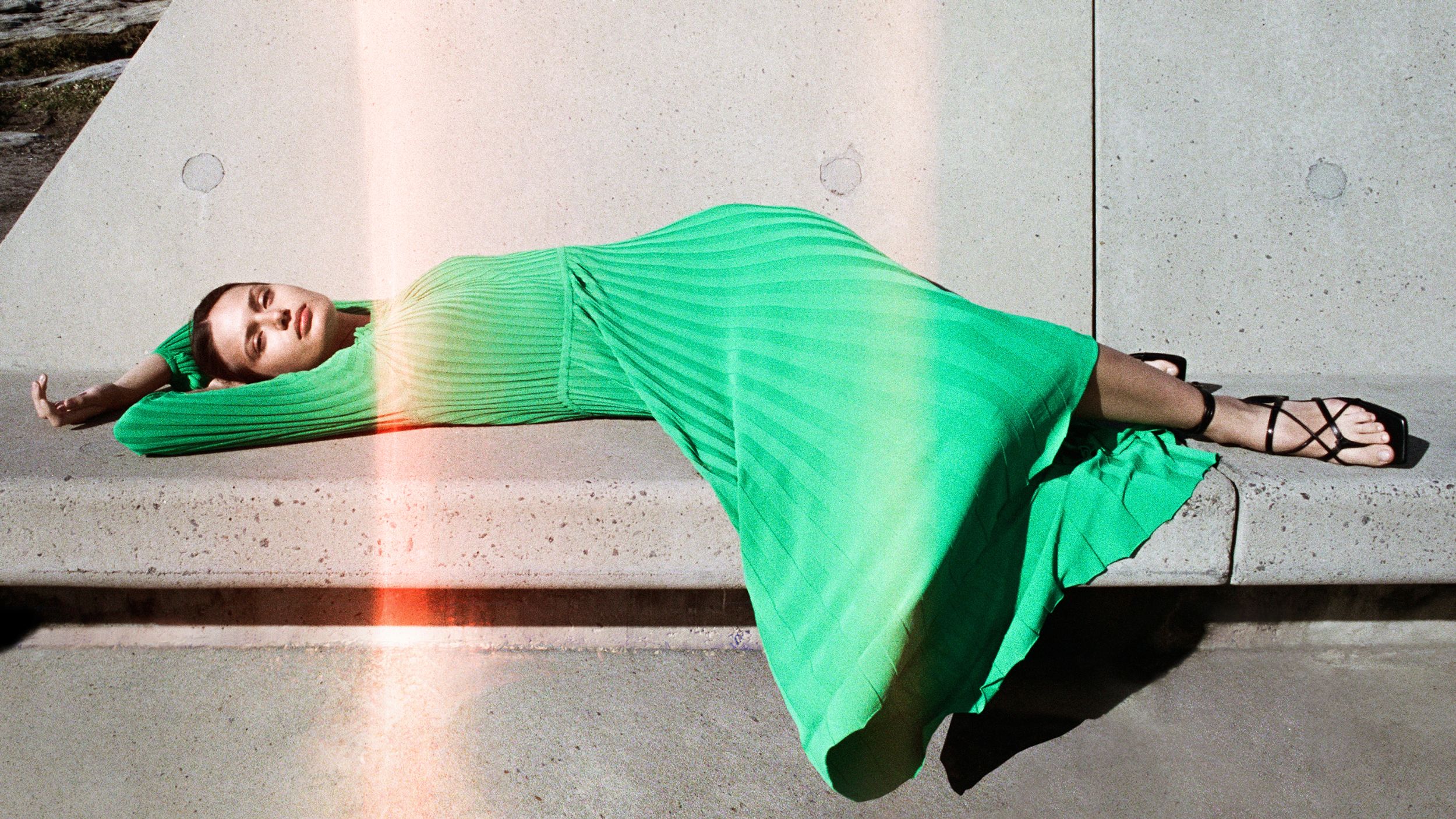 Brunette girl laying on concrete bench with her eyes closed, in green dress and black sandals.  