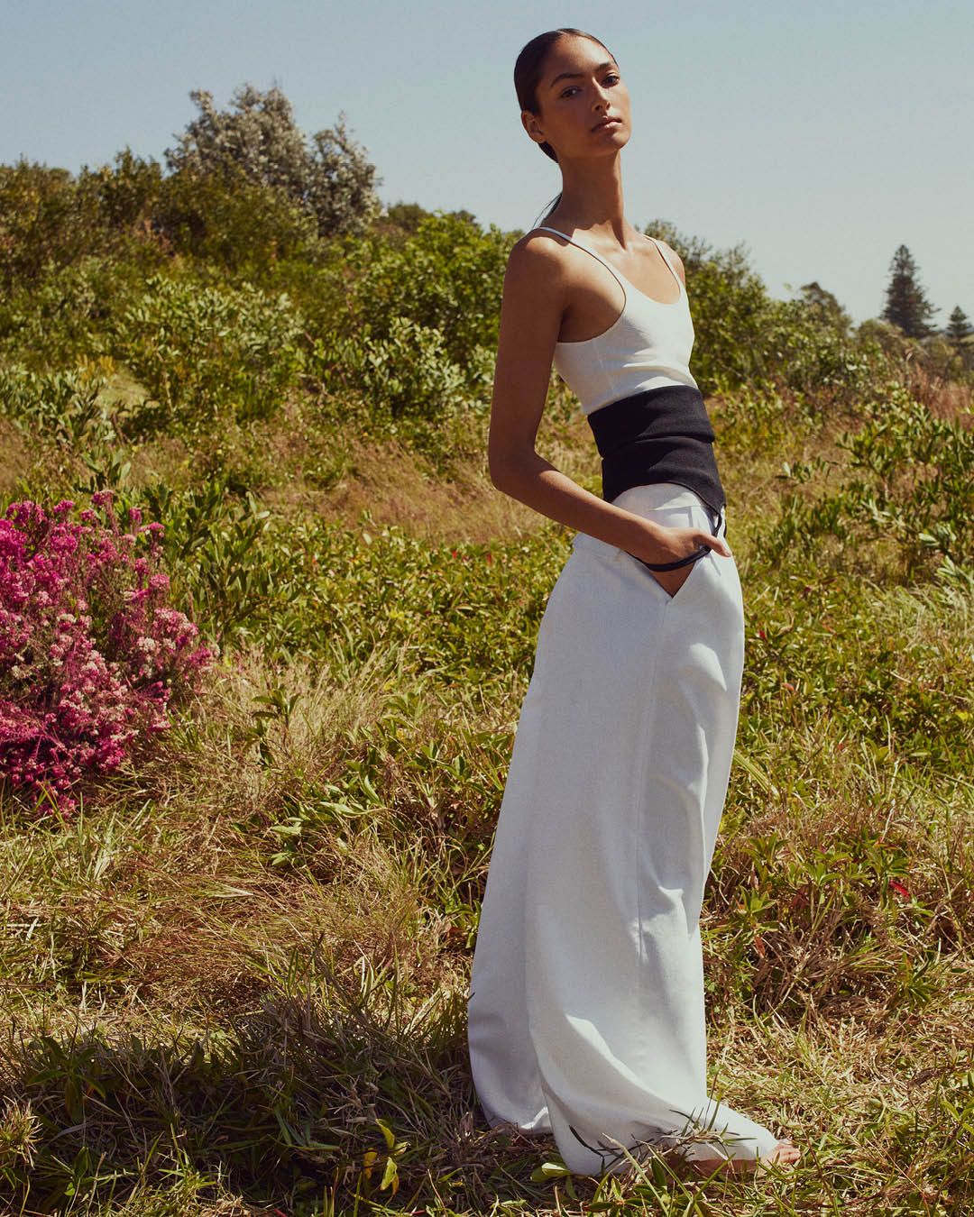Model standing on grass with hands in pockets wearing a white singlet and white wide-leg trousers