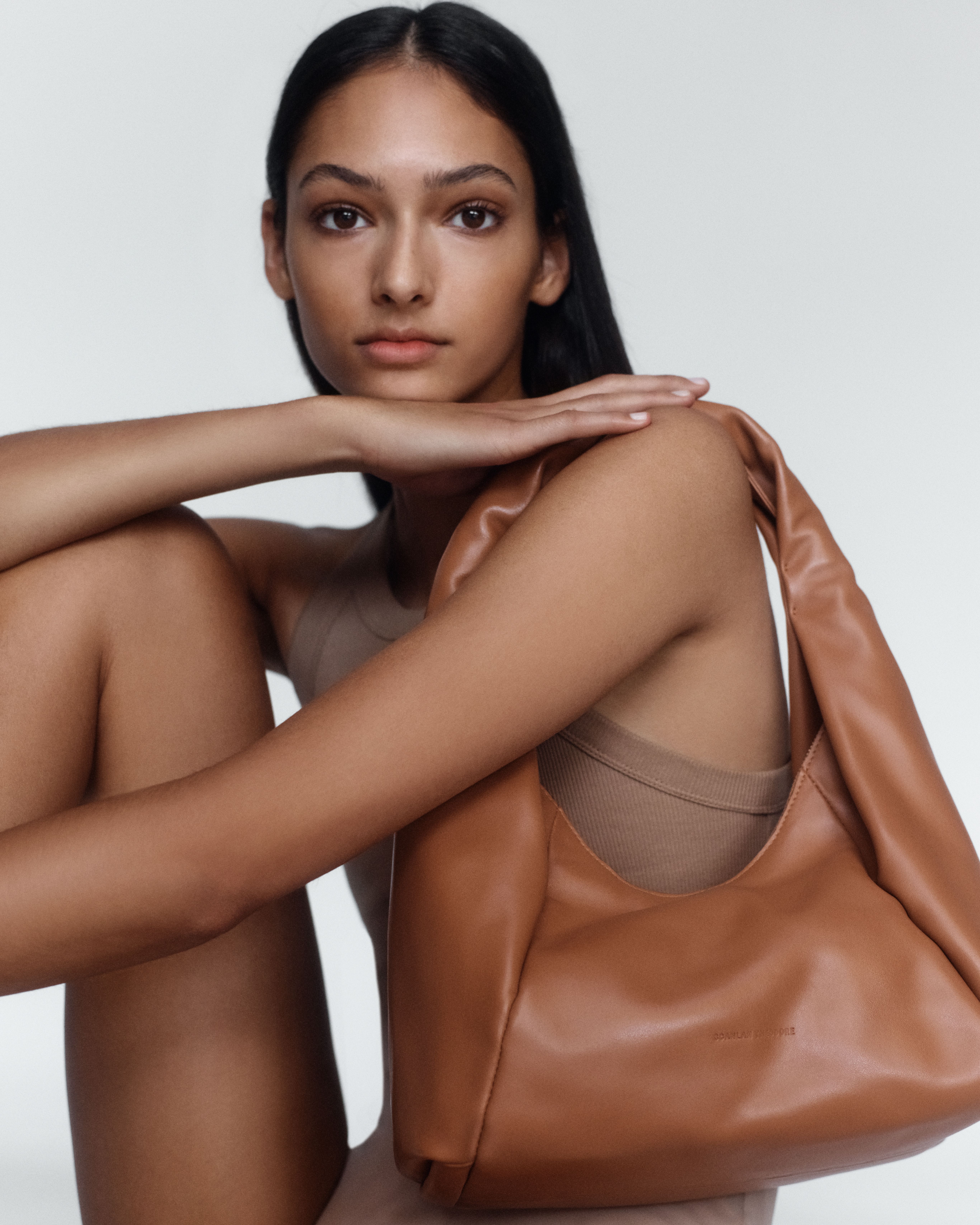 Model sitting with her arm resting on her knee and a tan leather shoulder bag on her shoulder