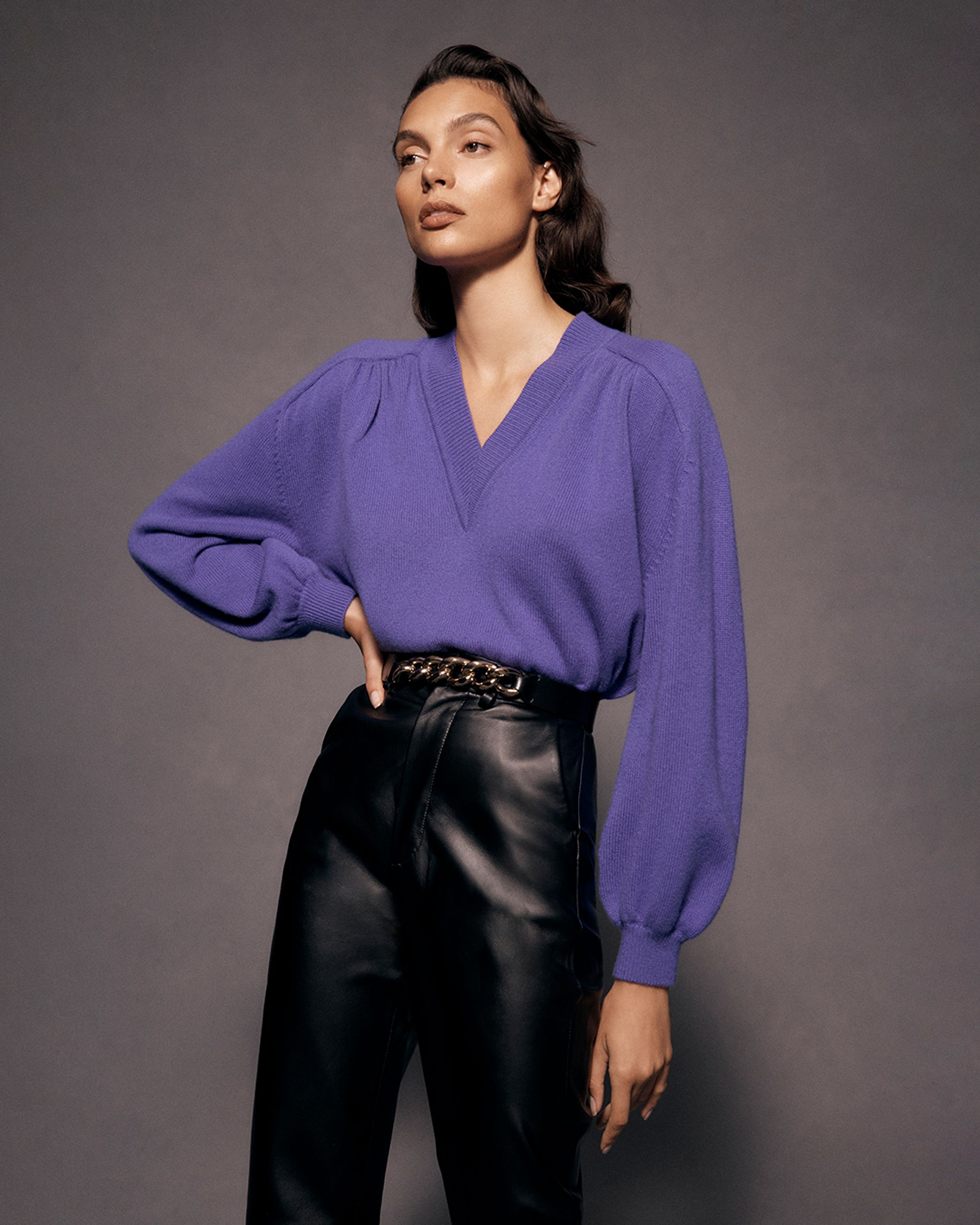 Brunette model posing with one hand on hips wearing a purple v-neck sweater and leather trousers