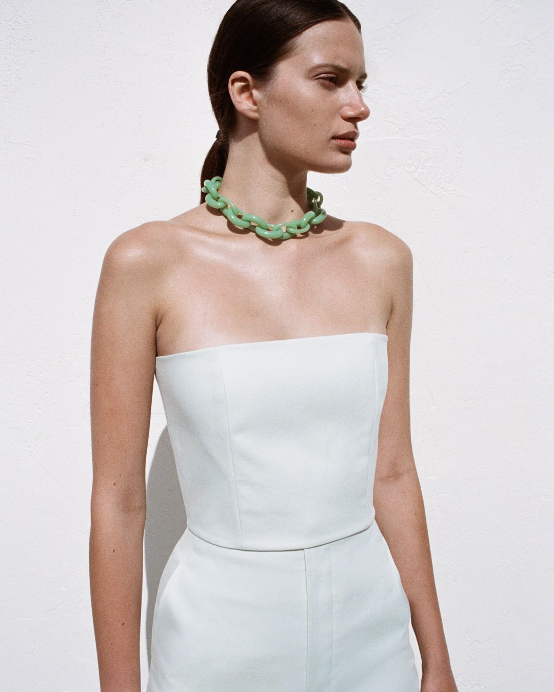 Brunette girl with ponytail wearing a green necklace and green strapless top, against white wall. 