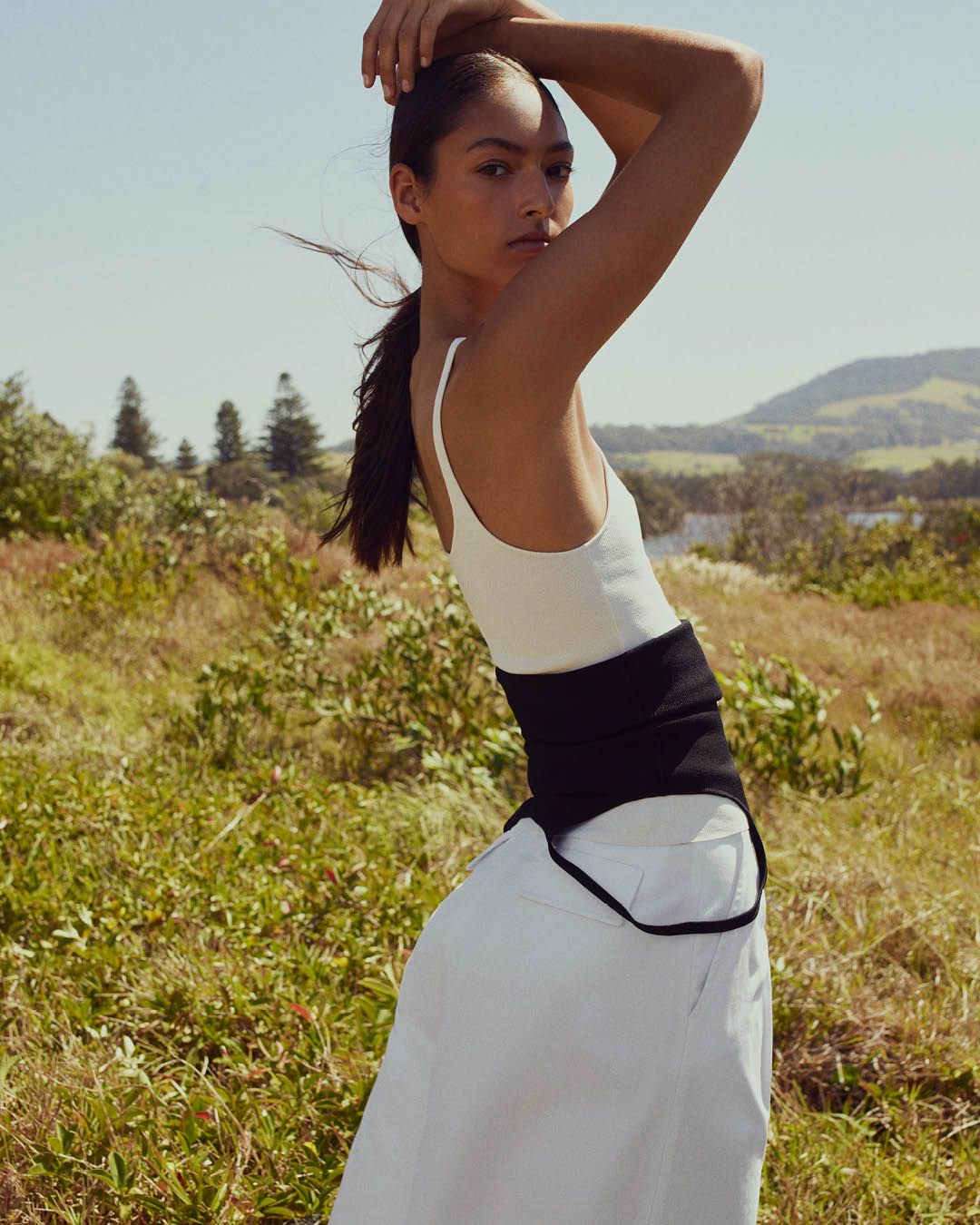 Model standing on grass with hands above her head wearing a white singlet and white wide-leg trousers