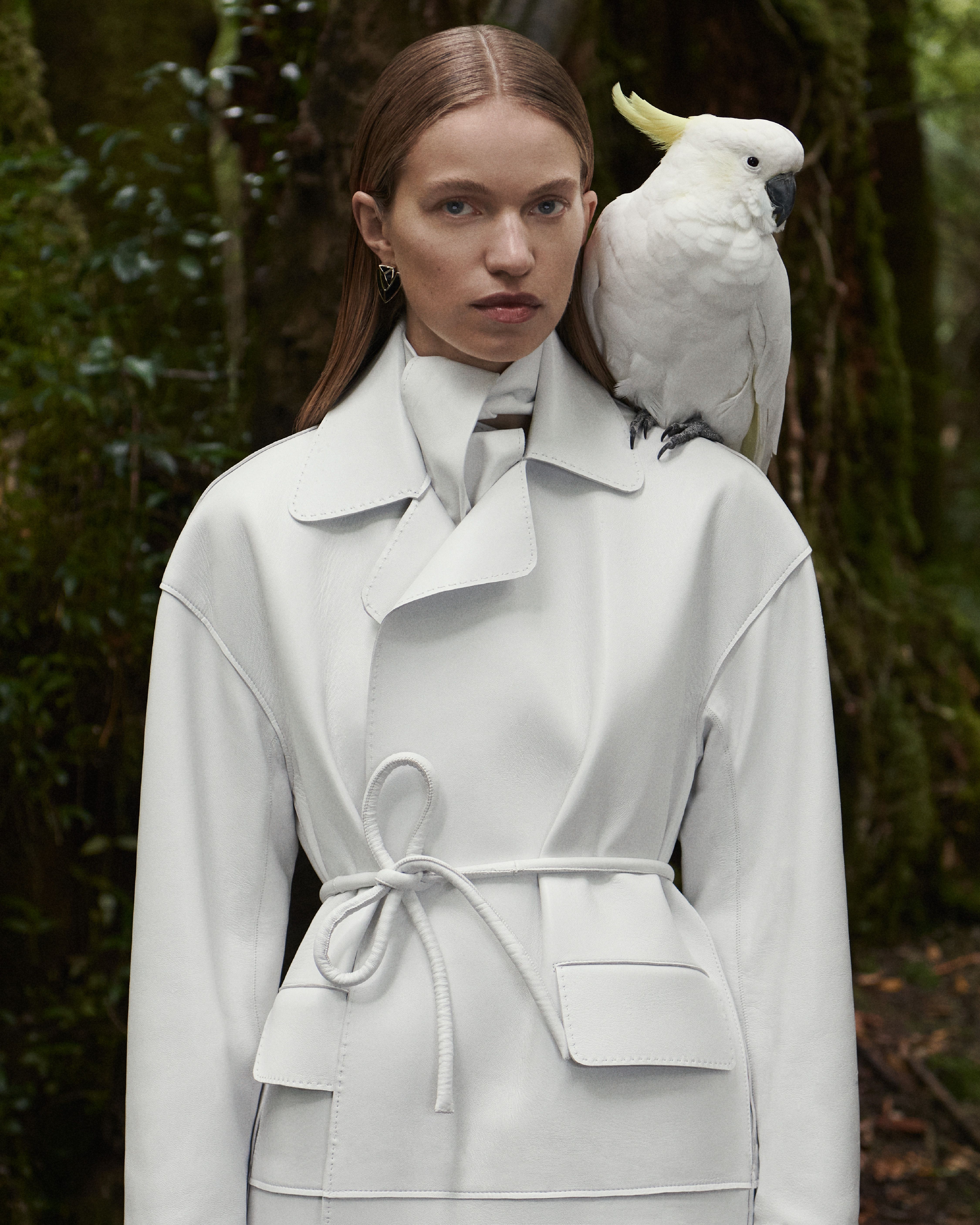 Sabine Glud standing in the forest wearing a white leather jacket with a cockatoo perched on her shoulder