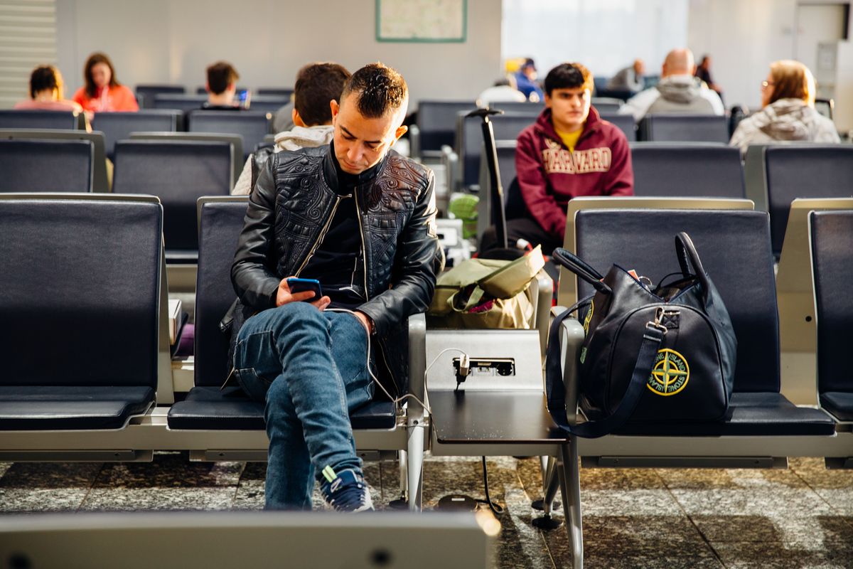 A traveler seated in a lounge chair charges his phone at a power outlet