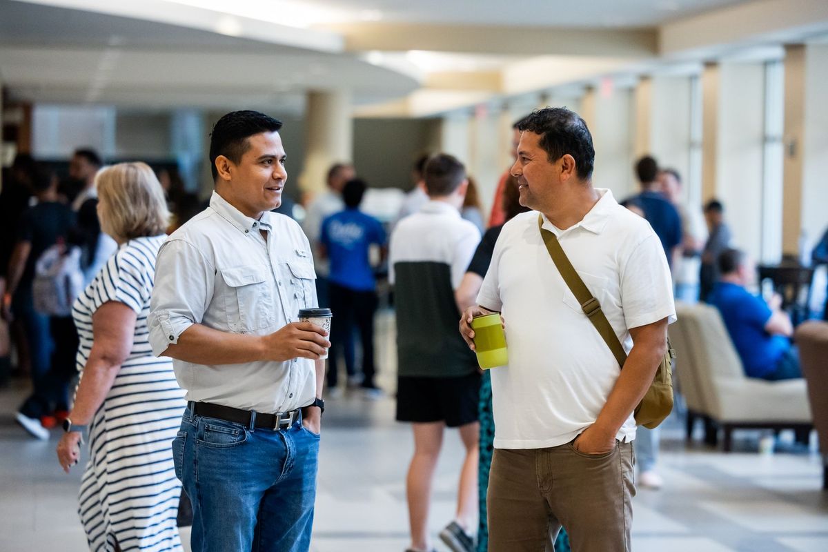 Two people standing in conversation at an indoor event, other attendees blurred behind them