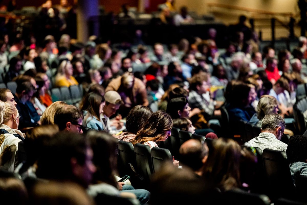 A warm-lit audience filling rows of seats at an indoor conference, the front row in focus