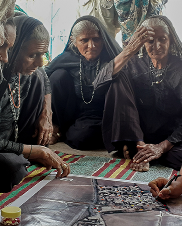 A group of women sit on the ground at a meeting and listen to a women speaking on the podium.