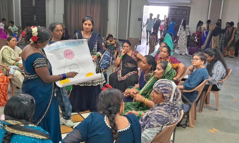 A woman in a blue saree is presenting a diagram on a chart to other women sitting in front of her