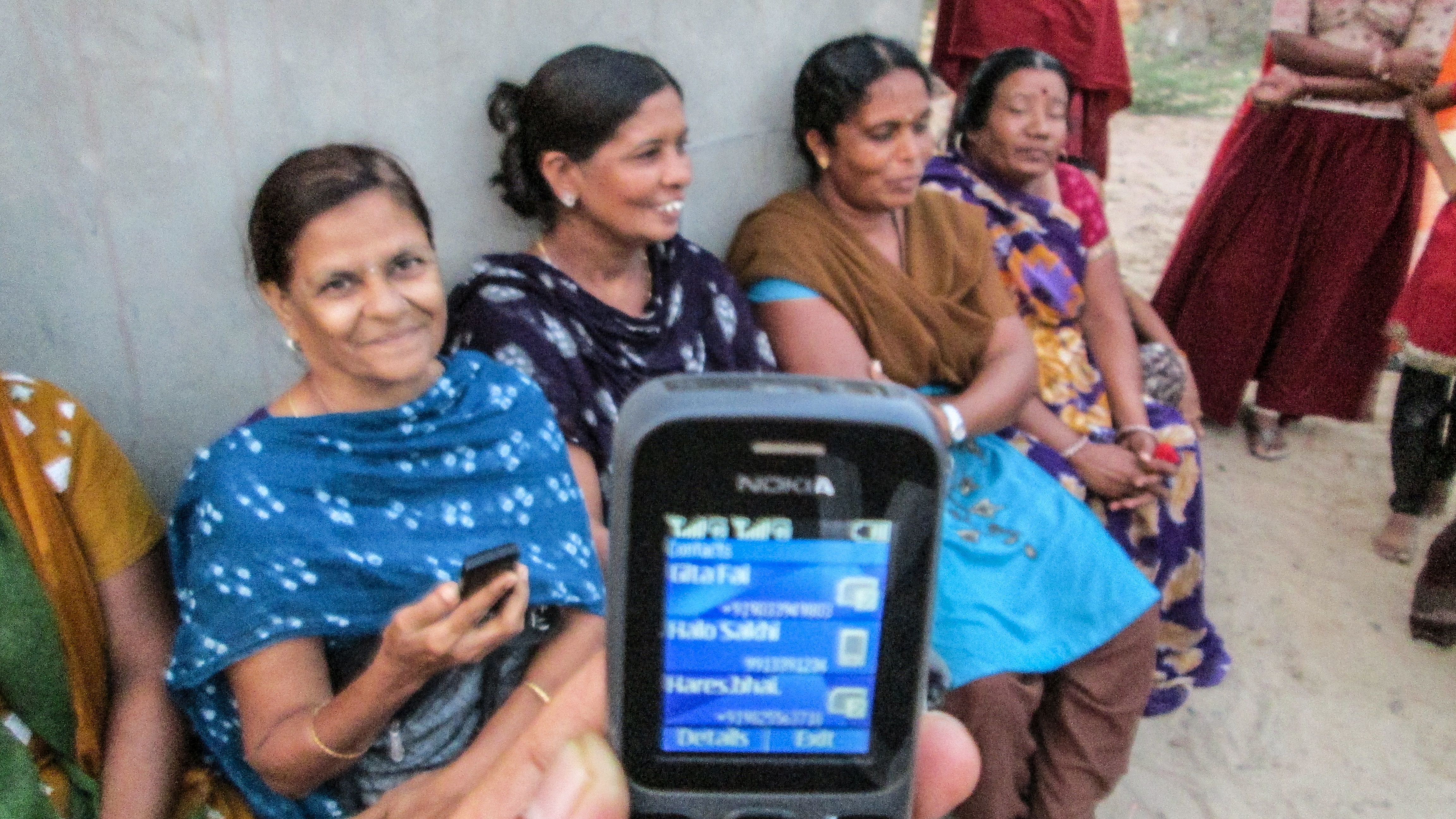 Four women are sitting on chairs and smiling and talking. There is a mobile phone screen in the foreground.