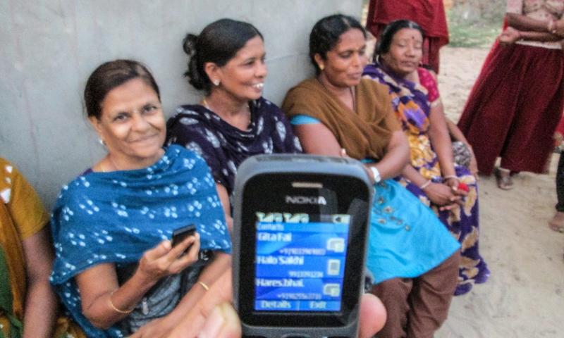 Four women are sitting on chairs and smiling and talking. There is a mobile phone screen in the foreground.