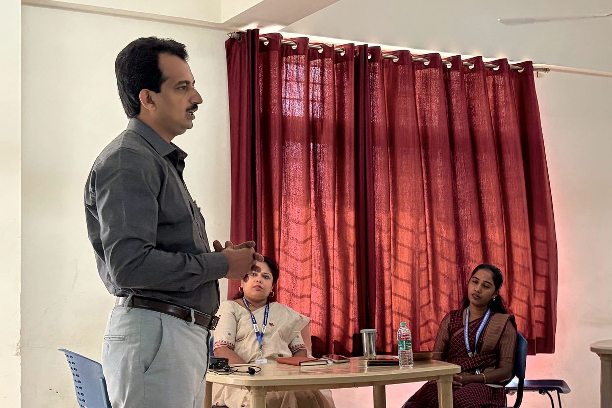 Dr. Manoj stands and addresses the room, with two participants seated at a table behind him.