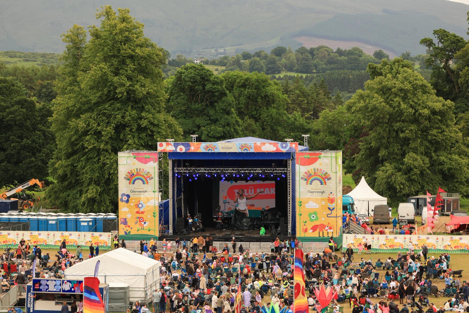 colourful main stage of a festival with crowds in foreground and trees and fields in background