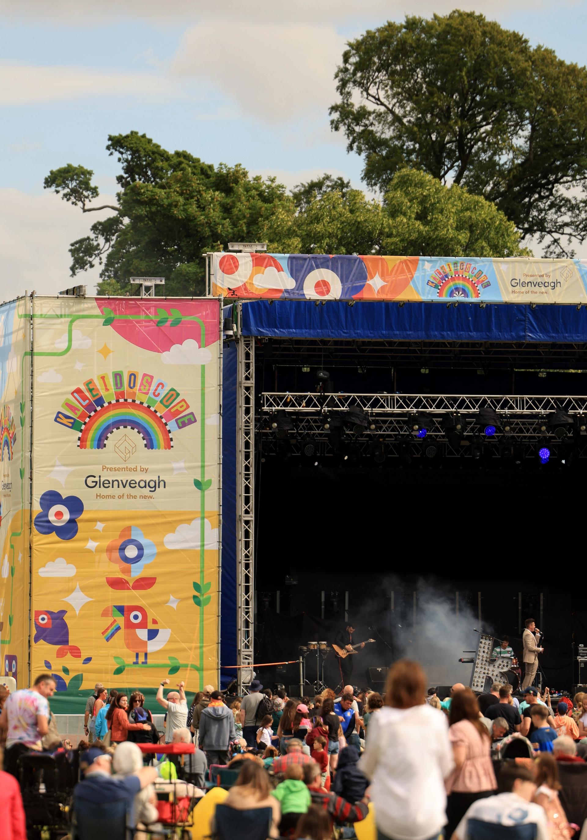 colourful main stage of a festival with crowds in foreground and trees and fields in background