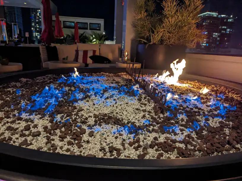 Fire pit with blue glass and flames at night on The Rooftop at Andaz San Diego, with lounge seating and city lights behind it.