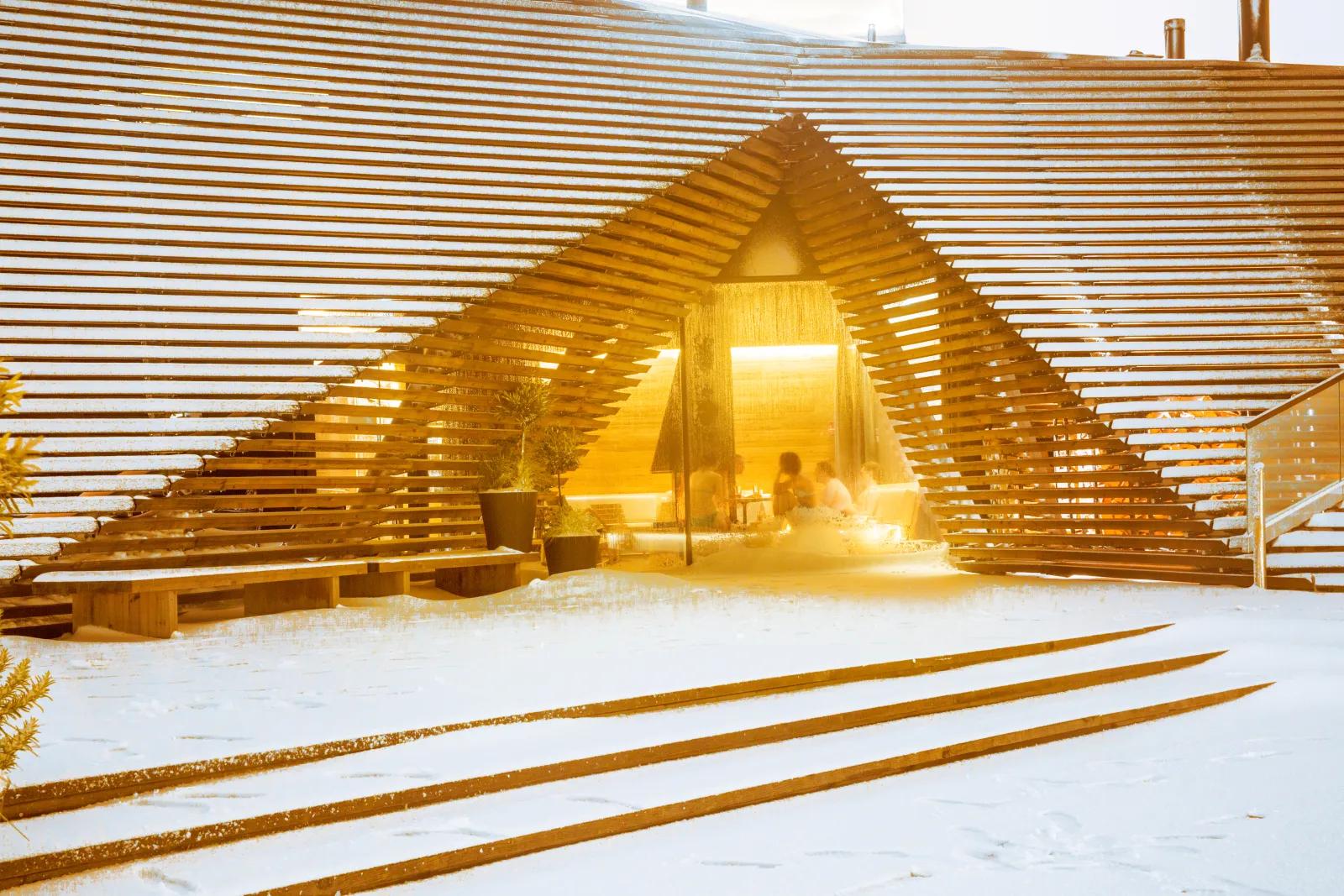 雪に覆われたLöylyの外観。木のラメラが三角形の開口部を縁取り、奥から夕日が差し込む