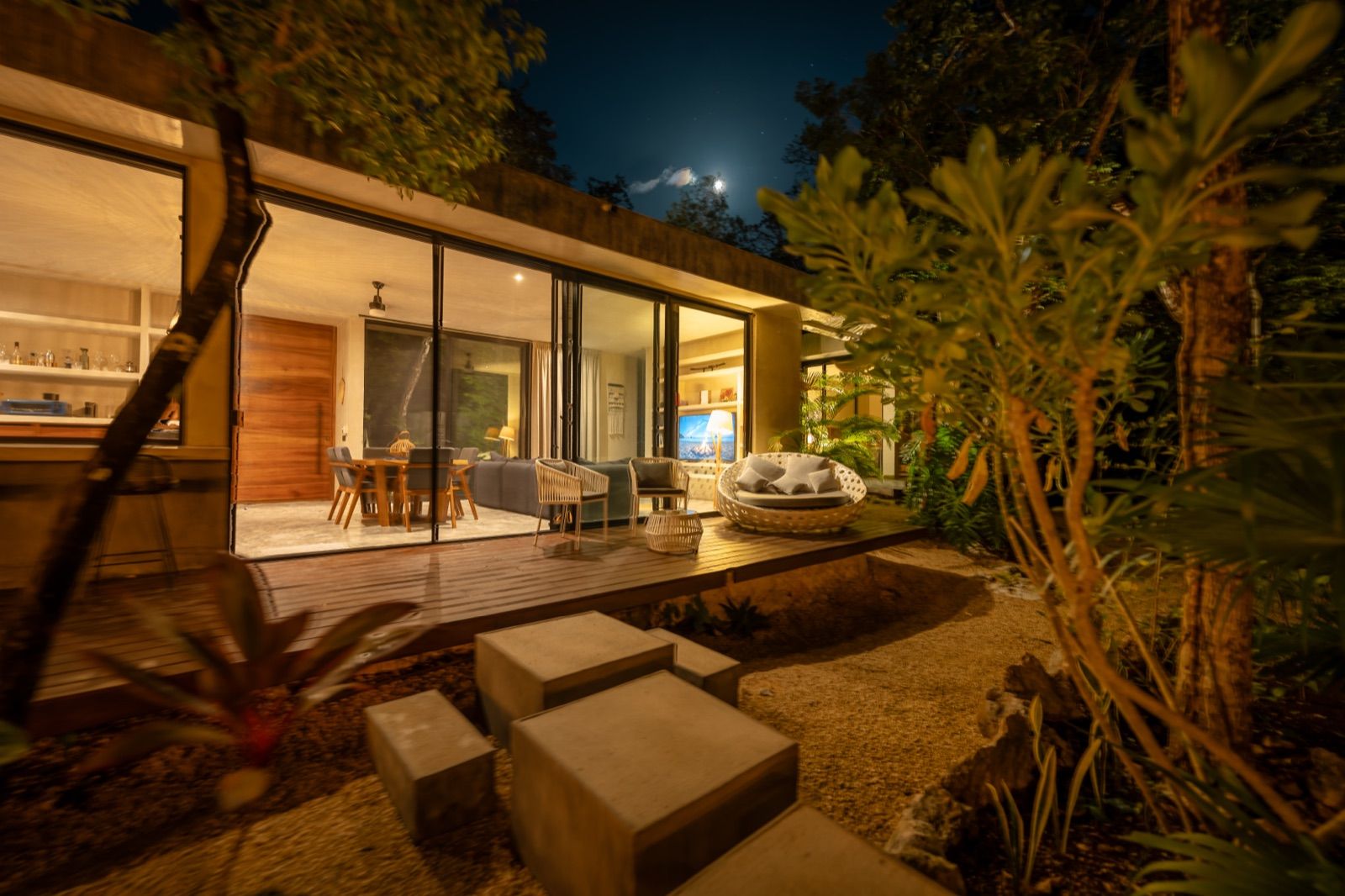 A glass-walled Kuyabeh residence lit up at night, deck and jungle under a moonlit sky