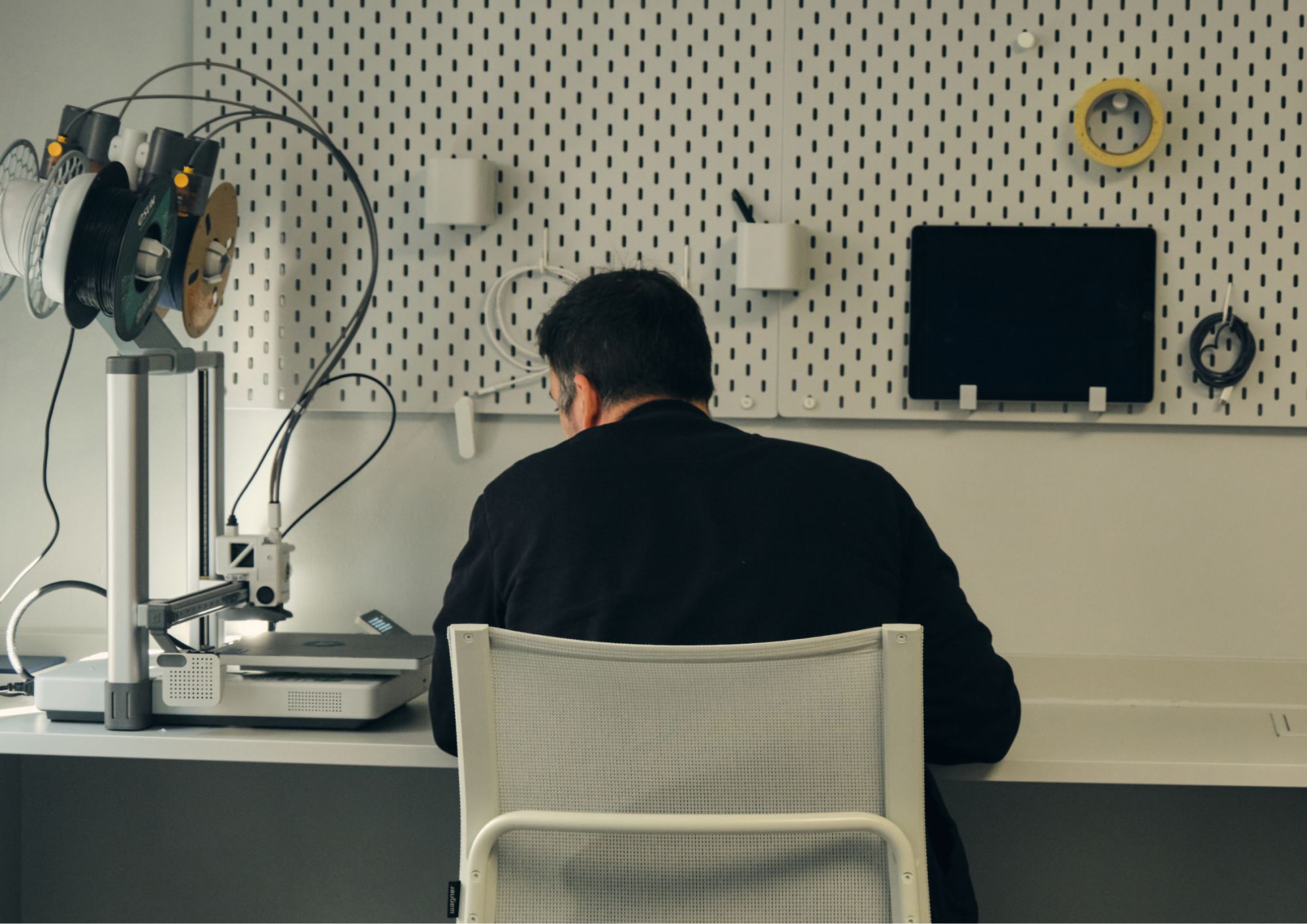 Person sitting at a workbench with a 3D printer and tools organized on a pegboard wall