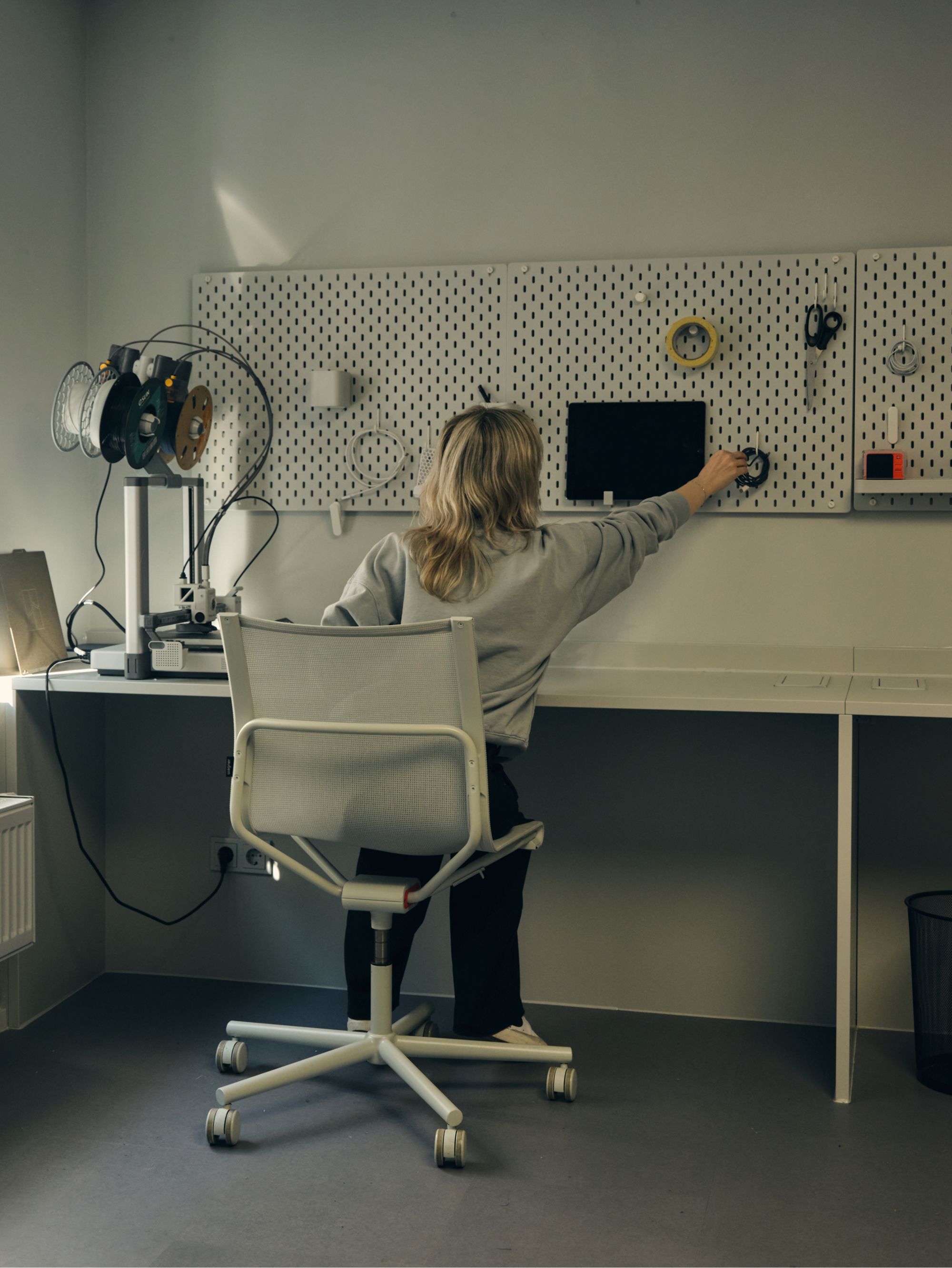 Person seated at a workbench, reaching for tools on a pegboard wall with a 3D printer nearby