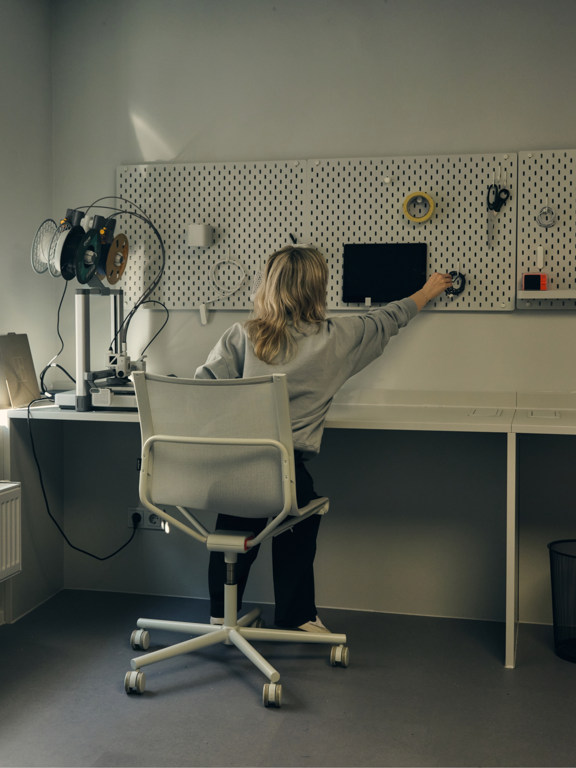 Person seated at a workbench, reaching for tools on a pegboard wall with a 3D printer nearby