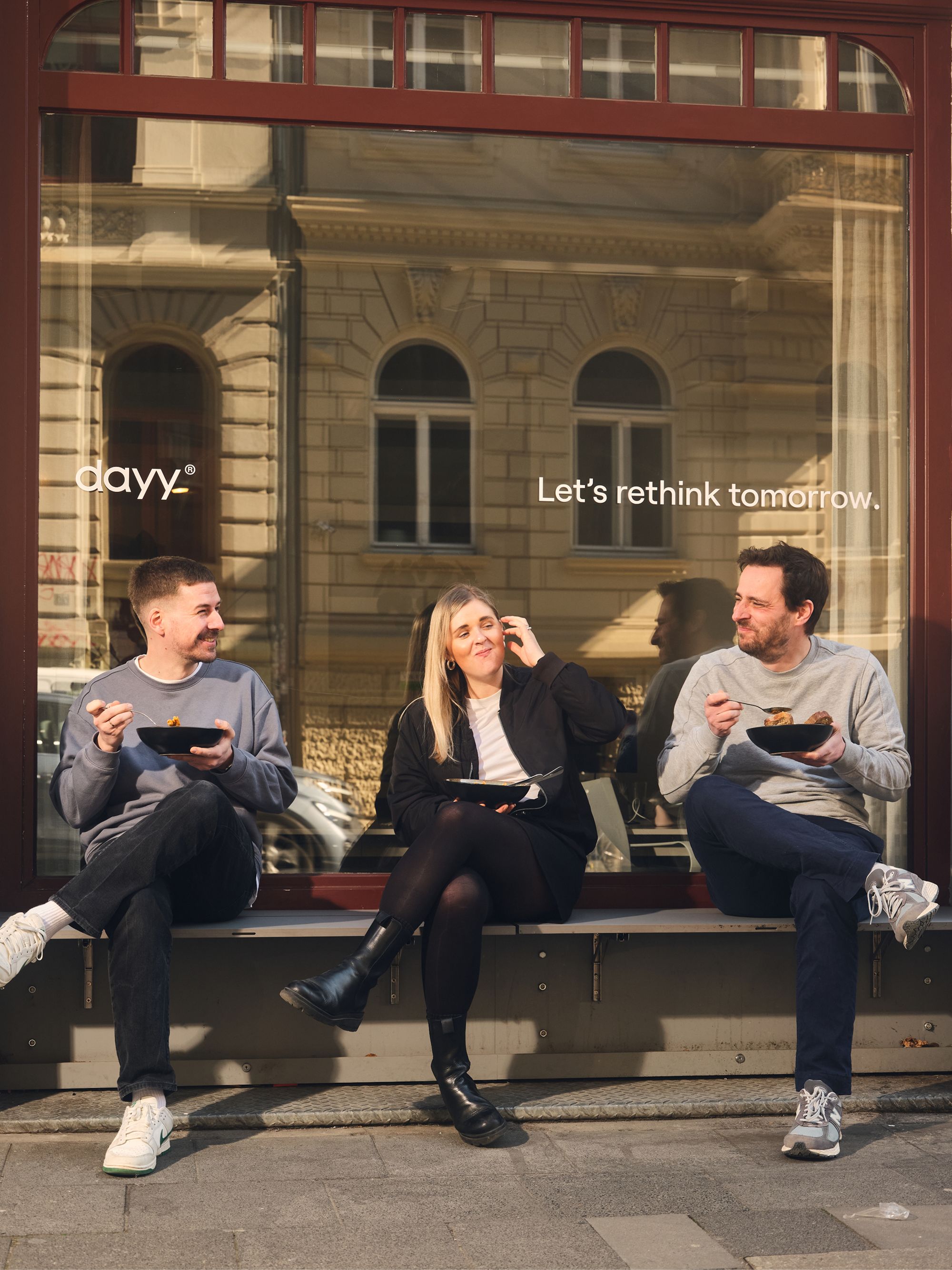Three people sitting outside on a bench, eating and talking in front of a large window with the text “Let’s rethink tomorrow”